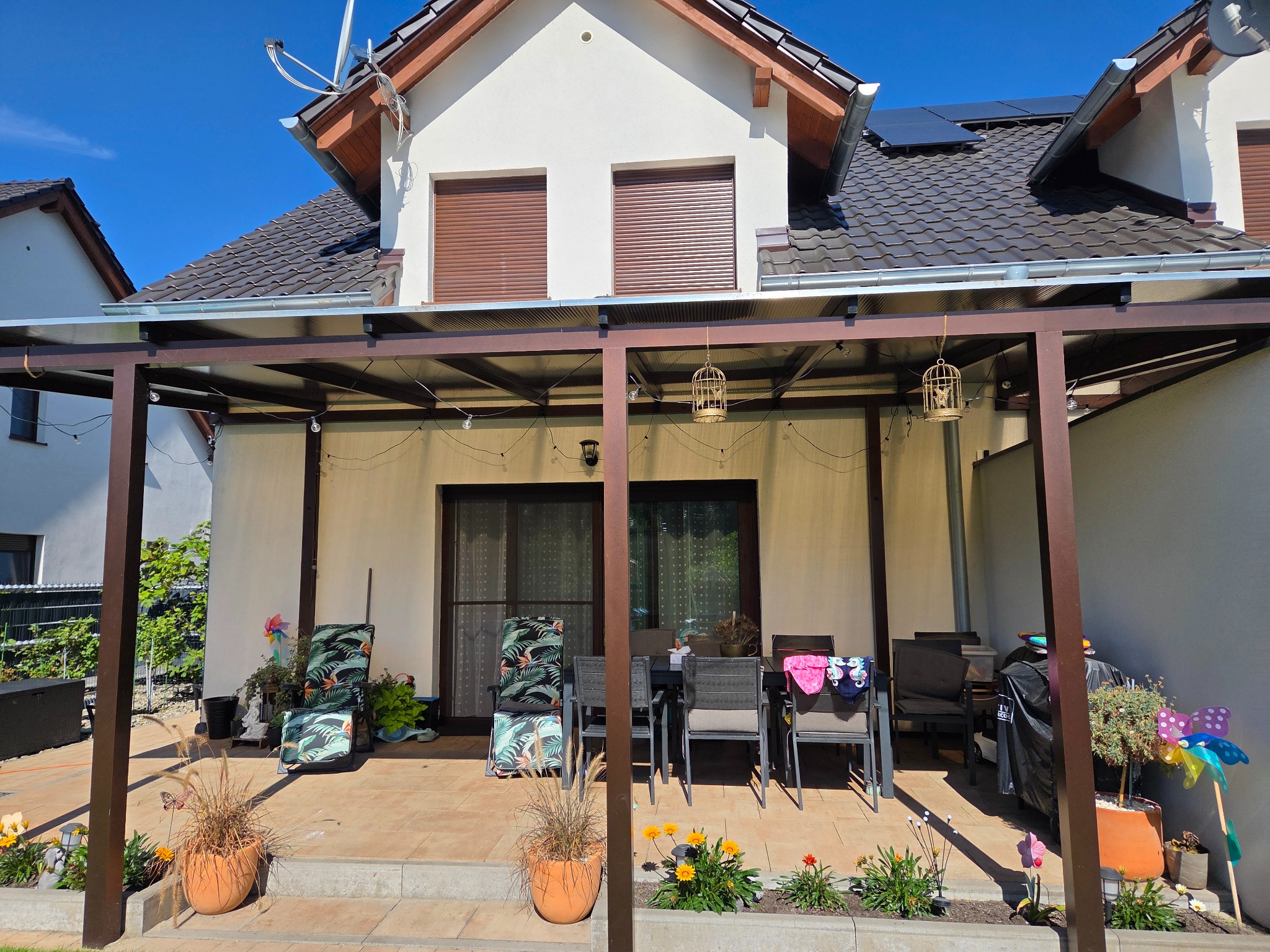 Zadaszony taras z brązową konstrukcją, stolikiem i krzesłami, kwiatami w donicach i dekoracjami. W tle dom z brązowymi roletami i panelami słonecznymi na dachu.