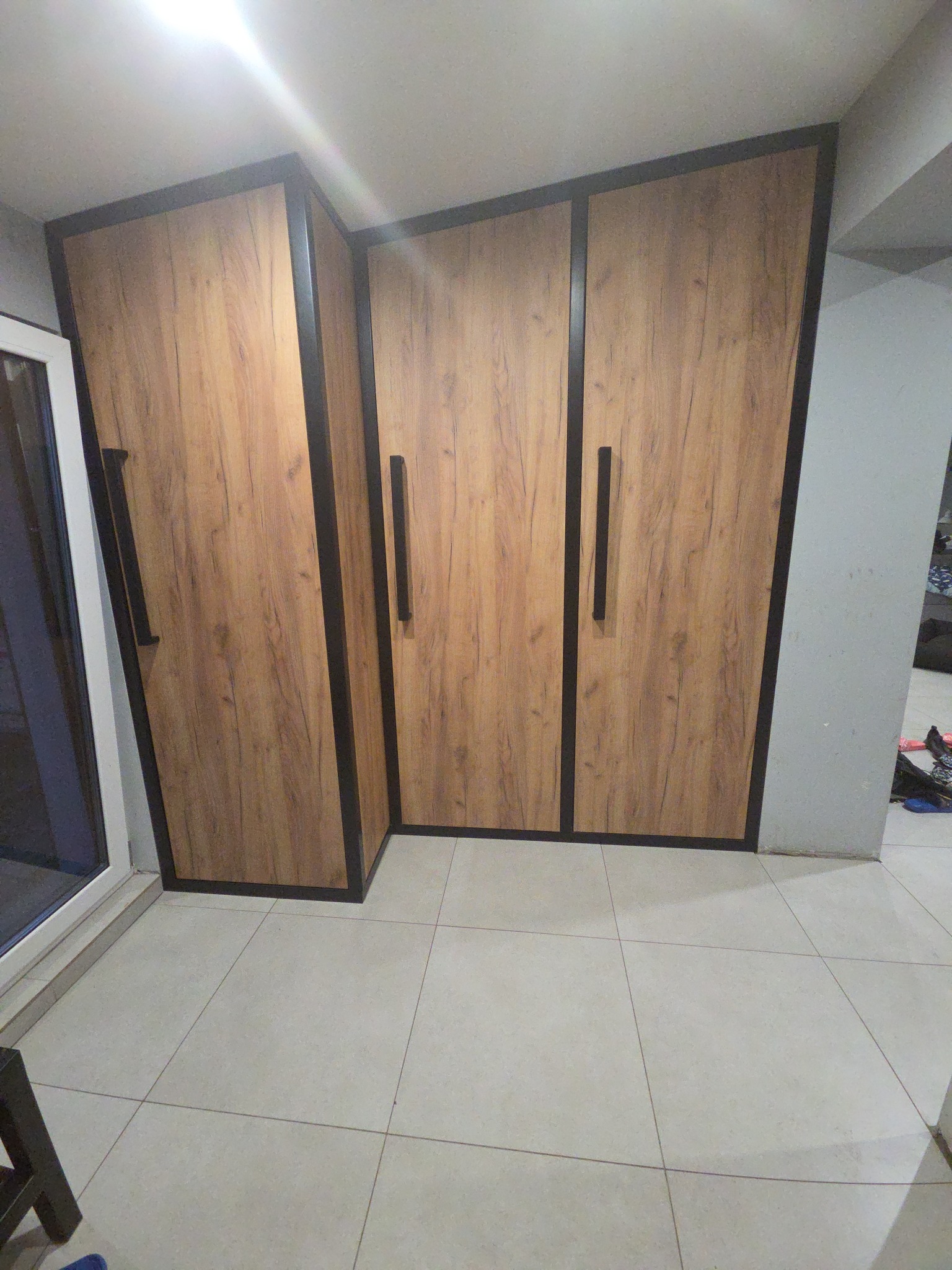 Trzydrzwiowa szafa z drewnopodobnymi frontami i czarnymi obramowaniami, widoczna w rogu pomieszczenia z jasną podłogą.