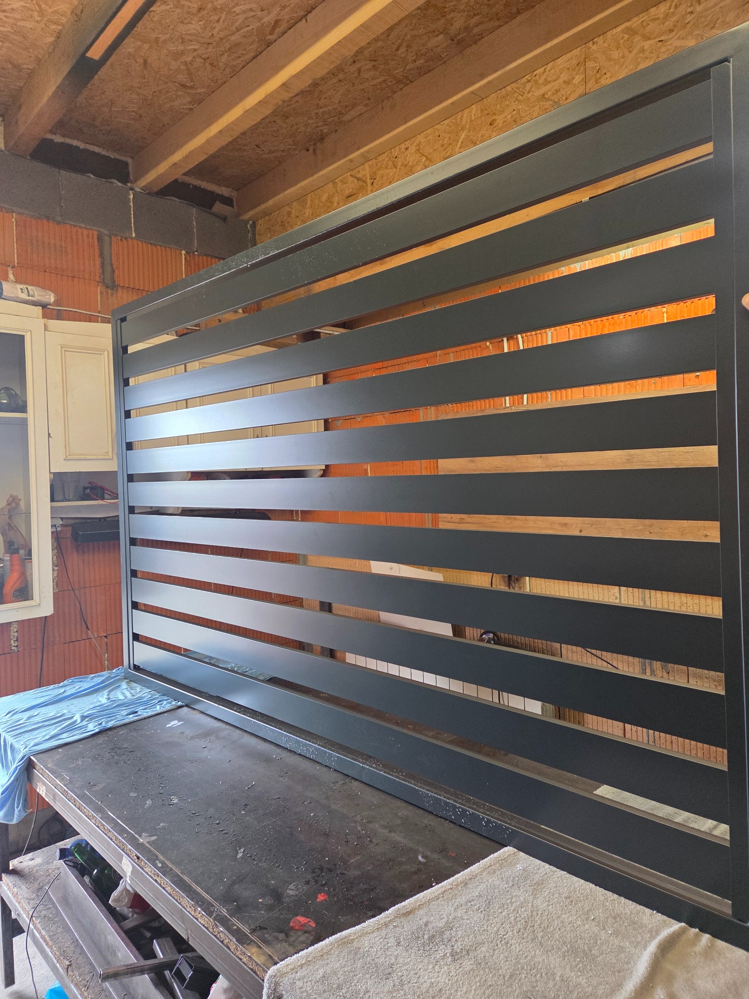 Częściowo zmontowana, nowoczesna brama wjazdowa z poziomymi panelami, w warsztacie. Widoczne cegły i elementy konstrukcyjne budynku w tle.