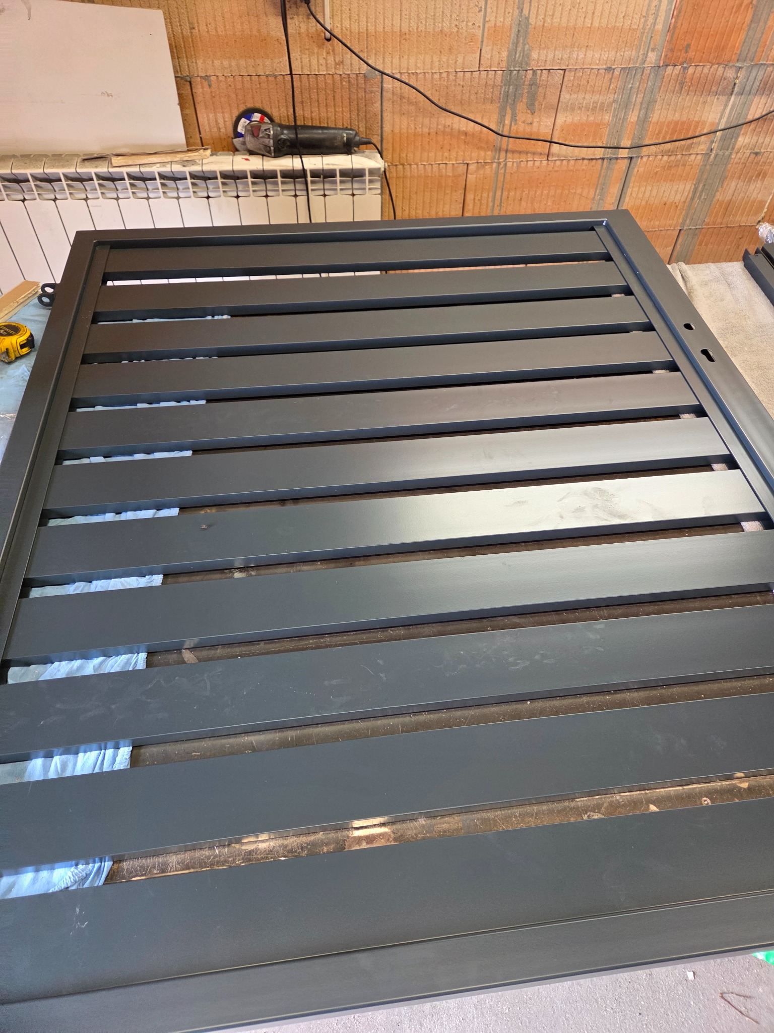 Szara, metalowa brama wjazdowa z poziomymi szczeblami, widoczna z bliska. W tle ceglana ściana i narzędzia.