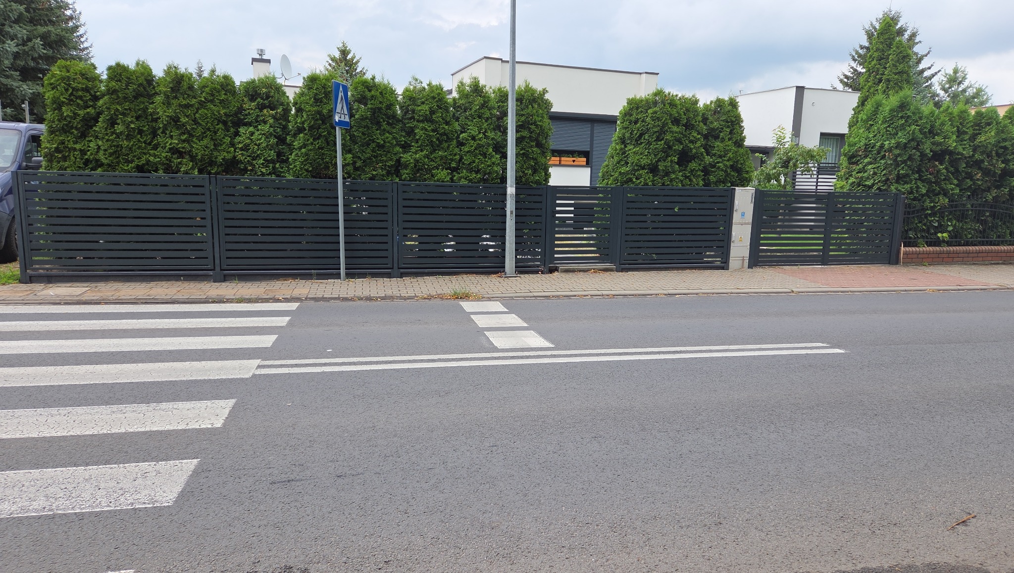 Nowoczesna, pozioma brama wjazdowa z metalowymi lamelami w kolorze antracytowym, widziana z perspektywy ulicy z przejściem dla pieszych i zielonymi tujami w tle.
