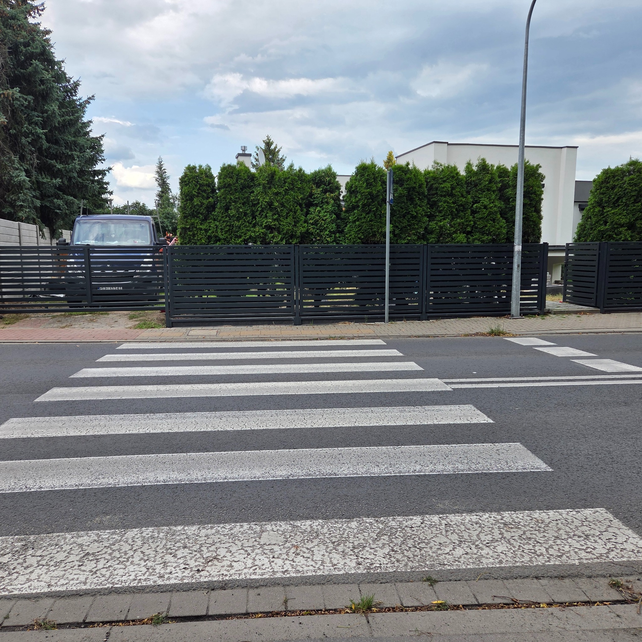 Nowoczesne, poziome ogrodzenie metalowe w kolorze grafitowym, widoczne z poziomu przejścia dla pieszych, z zielonymi tujami w tle i fragmentem zaparkowanego auta.