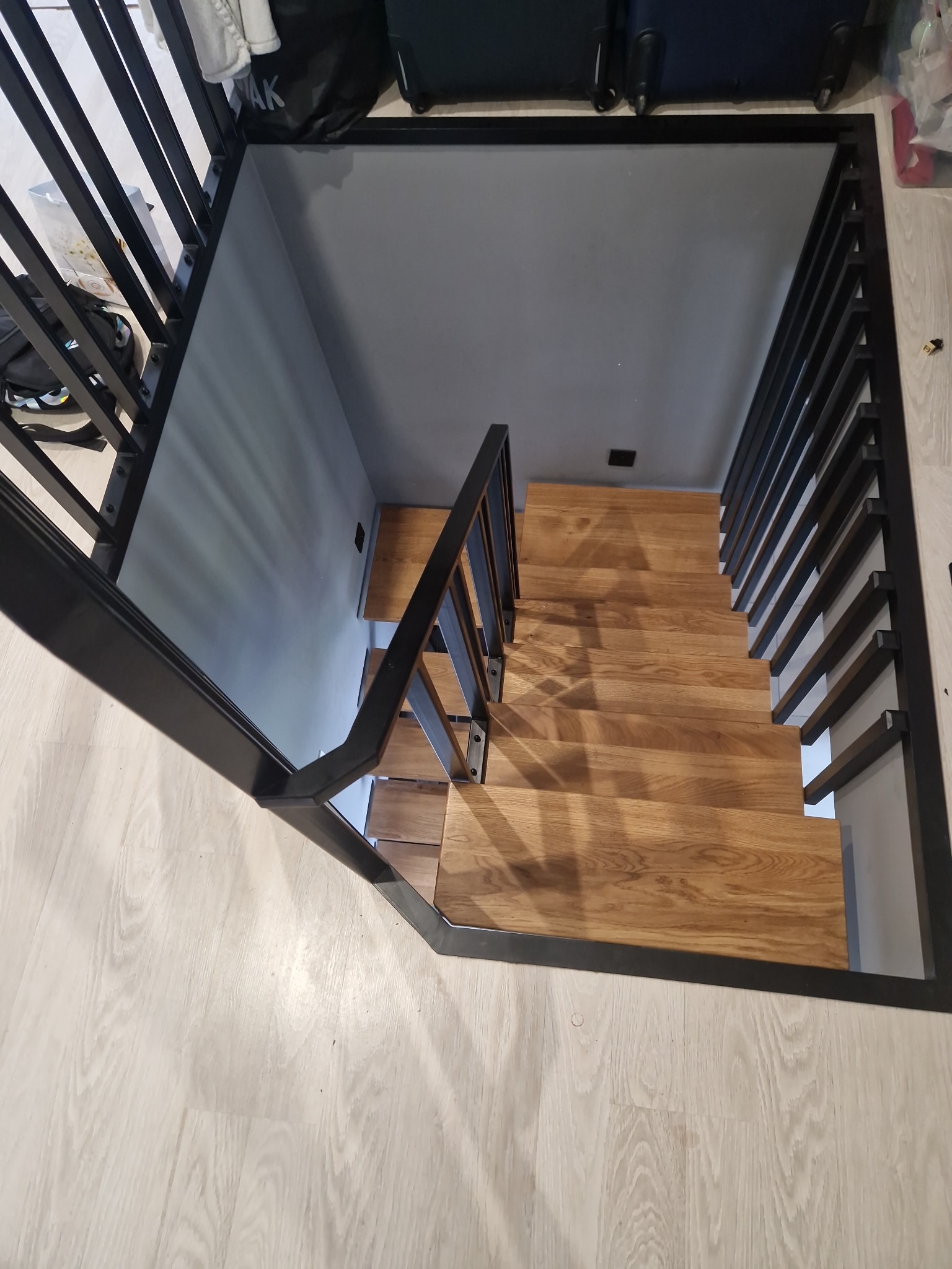 Drewniane schody z czarną, metalową balustradą, ujęte z góry. Widoczne schody prowadzą w dół, oświetlone światłem naturalnym. Minimalistyczny design.