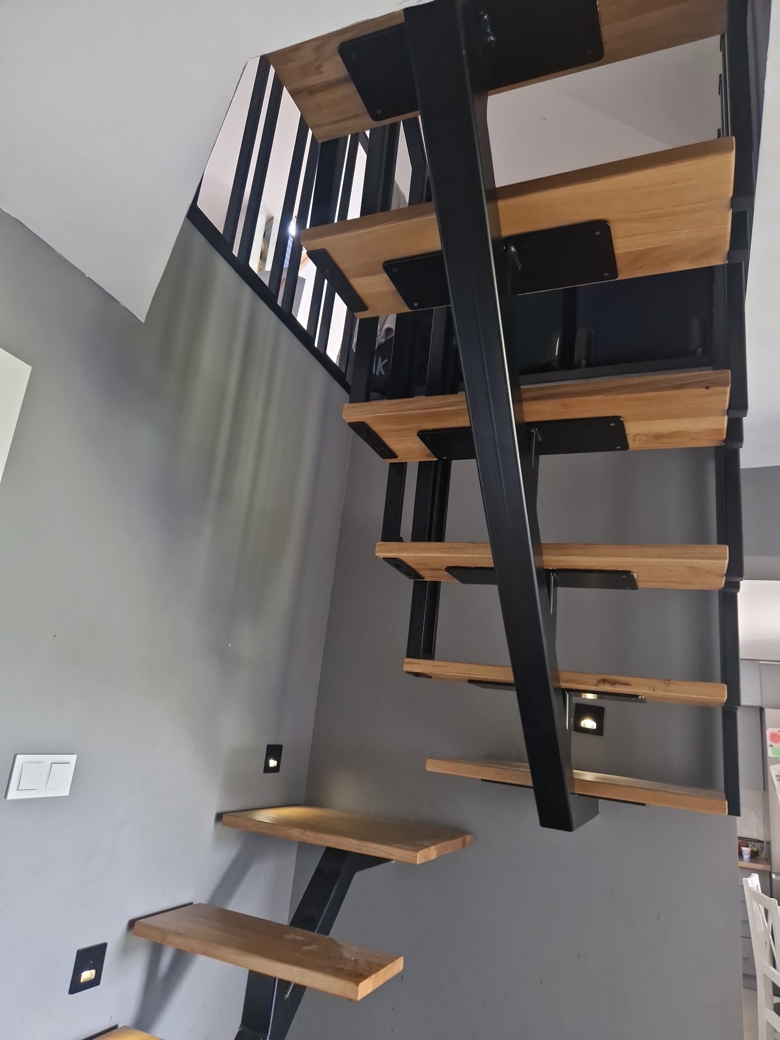 Nowoczesne schody drewniane z metalową konstrukcją, zamontowane przy szarej ścianie z wbudowanym oświetleniem. Minimalistyczny design.