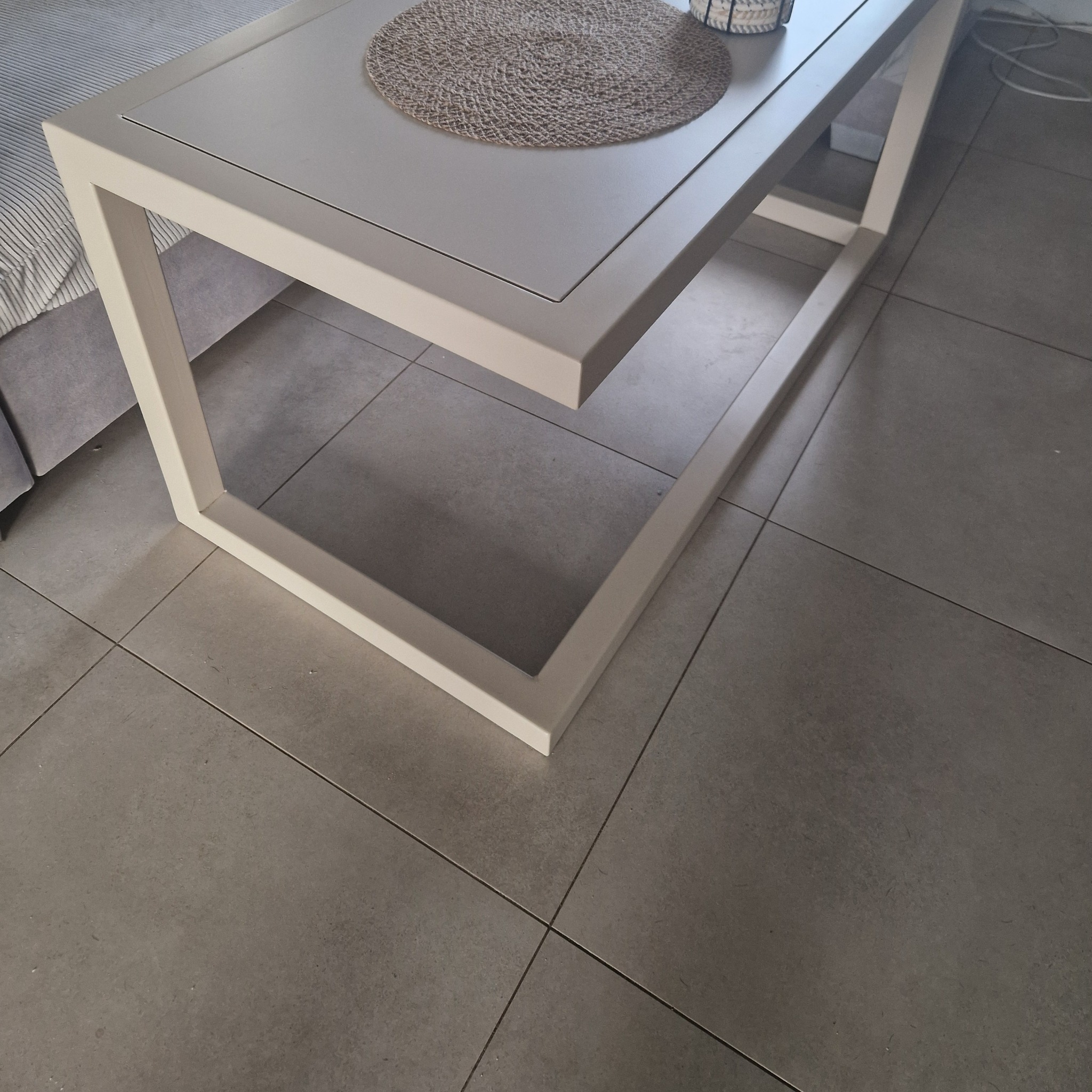 Stolik kawowy w stylu minimalistycznym z jasnym blatem i geometryczną podstawą. Na blacie leży okrągła podkładka. Widoczna szara podłoga z płytek.