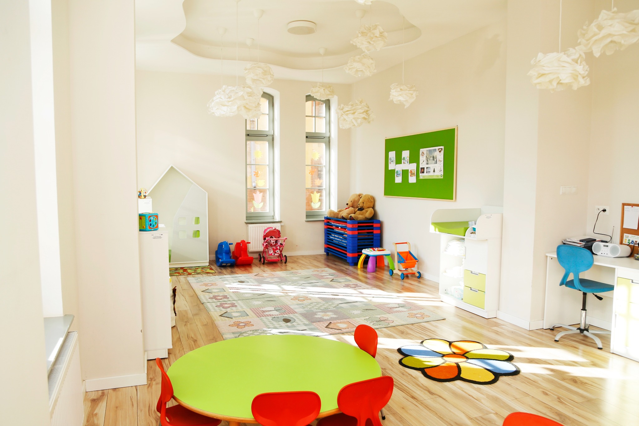 Jasne wnętrze sali zabaw z okrągłym, zielonym stolikiem i czerwonymi krzesłami. W tle dywan w kwiaty, zabawki, biurko z krzesłem i dekoracje w kształcie chmur.