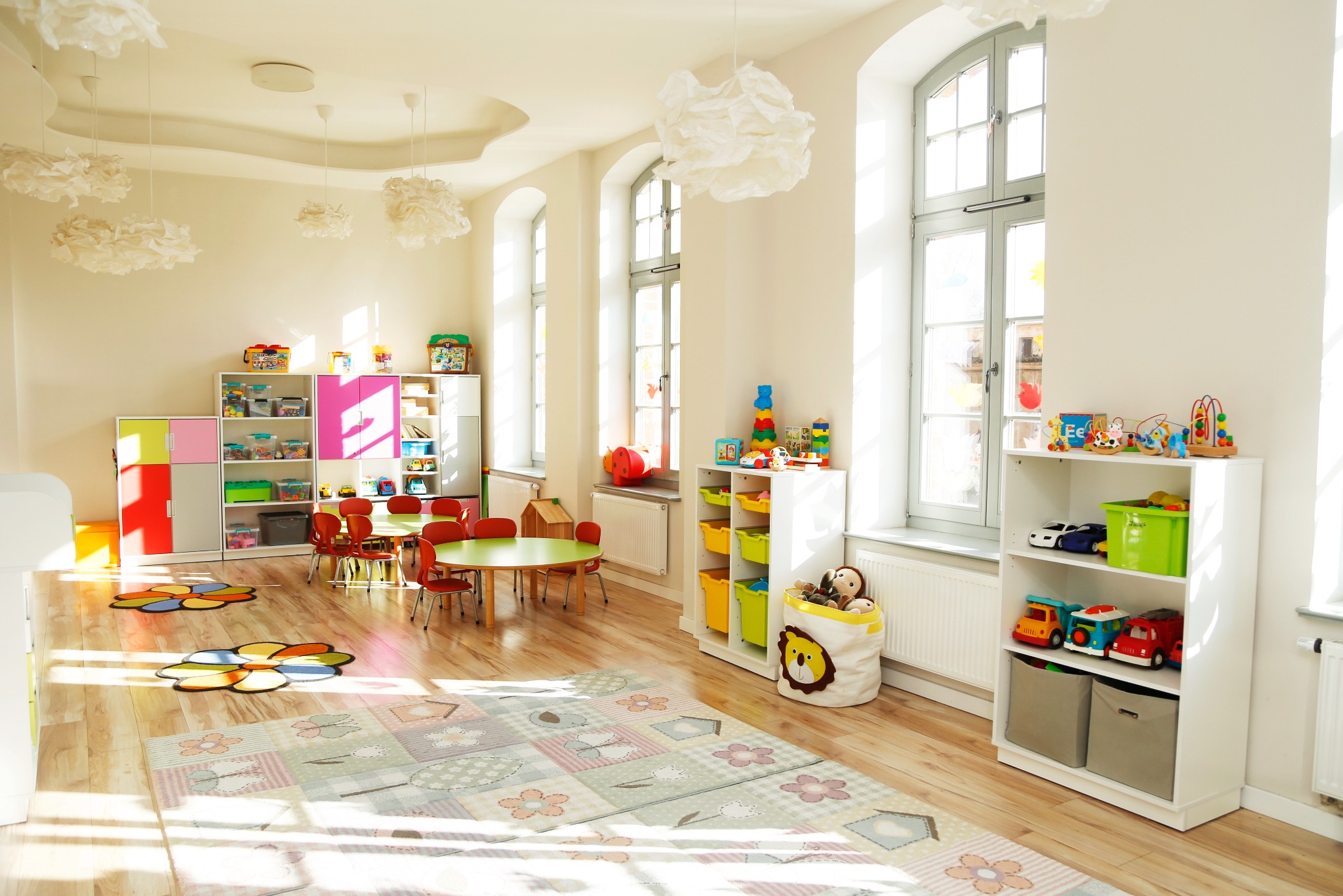 Jasny, przestronny pokój zabaw w żłobku z drewnianą podłogą, kolorowymi meblami, zabawkami i dywanem w pastelowe wzory. Duże okna wpuszczają naturalne światło.