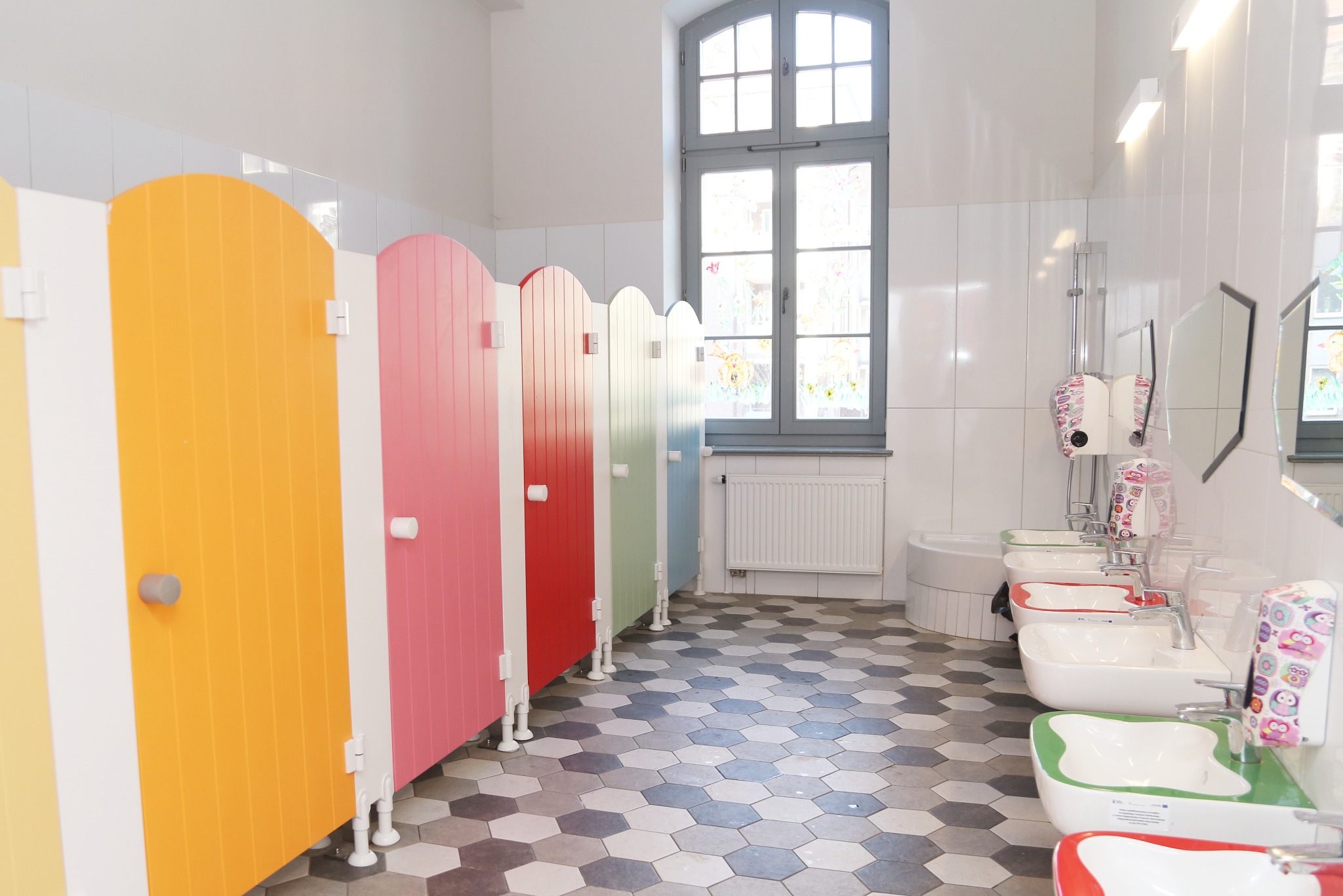 Kolorowe kabiny i umywalki dla dzieci w łazience. Żółta, różowa, czerwona i zielona kolorystyka. Heksagonalna podłoga. Okno z dekoracjami.