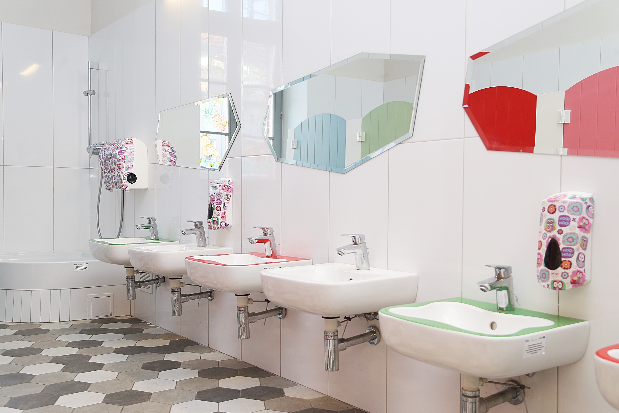 Umywalki dla dzieci w żłobku z kolorowymi obwódkami (zielona, czerwona) i lustrami o nieregularnych kształtach. Dozowniki mydła w sówki. Płytki heksagonalne na podłodze.