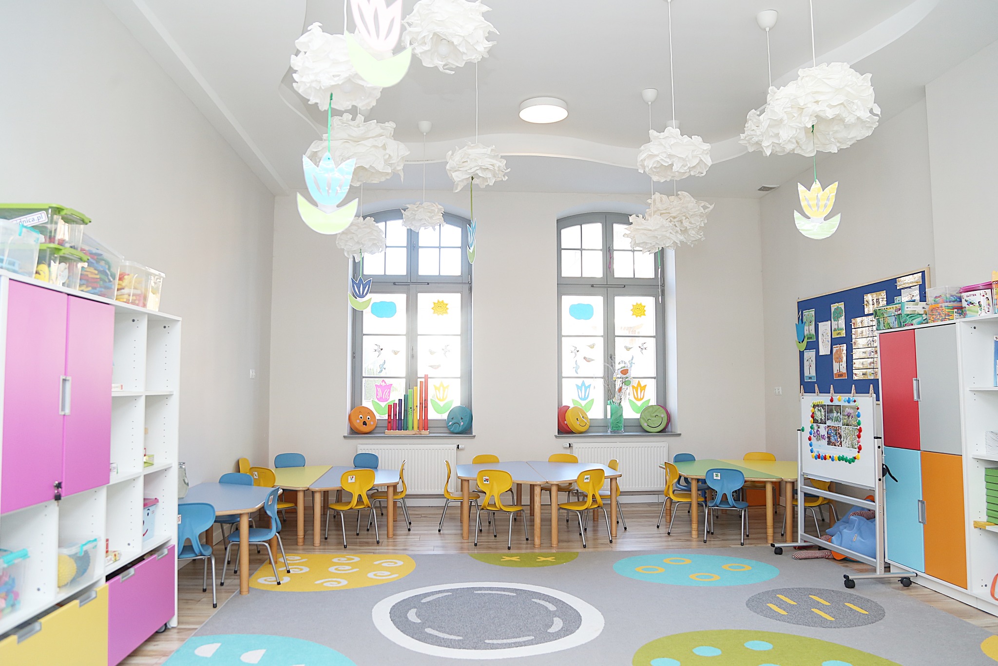 Jasna sala żłobka z kolorowymi meblami i dekoracjami: stoliki z krzesłami, regały z zabawkami i dywan w kropki. Okna zdobią naklejki z motywami roślinnymi.