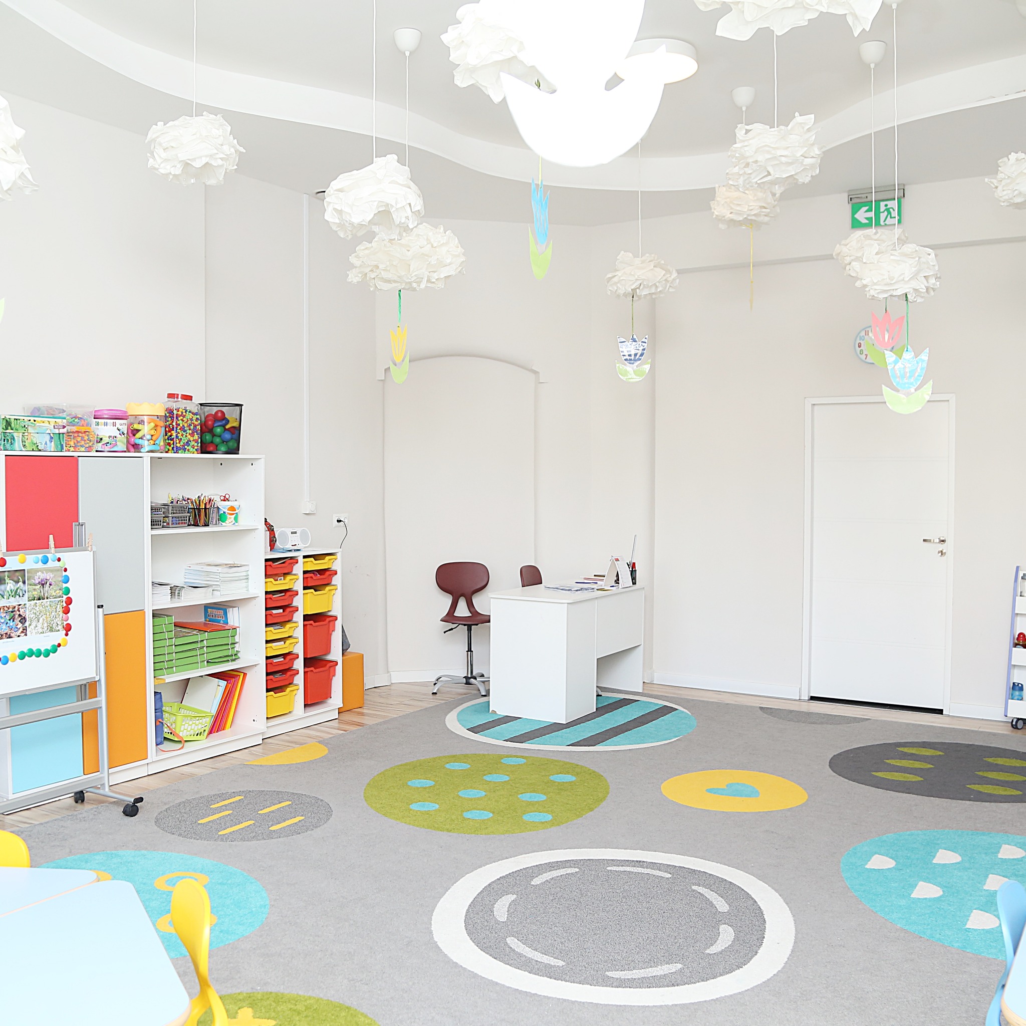 Jasne wnętrze sali zabaw z kolorowym dywanem w okręgi, białymi meblami i dekoracjami sufitowymi przypominającymi chmury i kwiaty, tworząc przyjazną przestrzeń.
