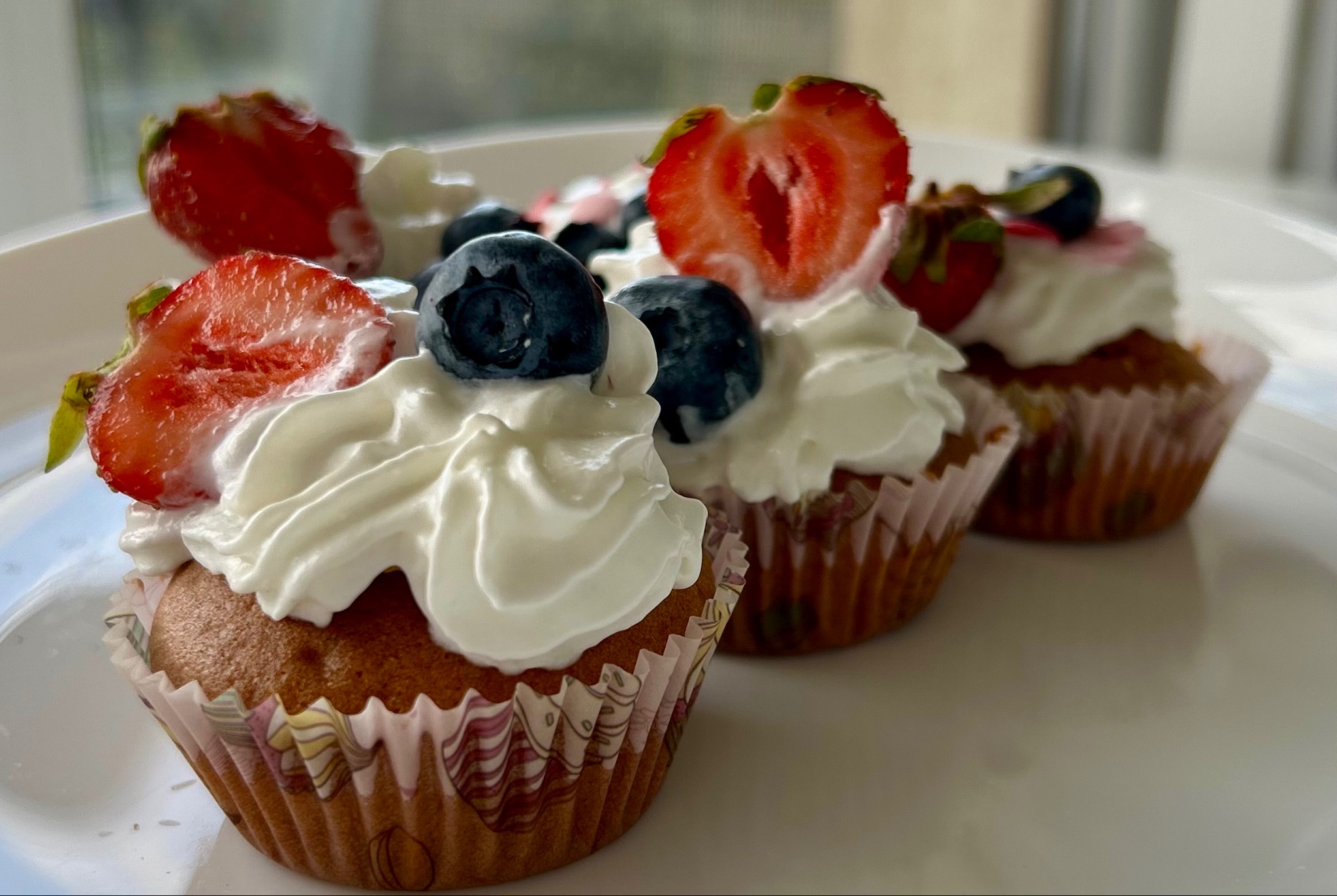 Rząd czterech apetycznych babeczek z kremem i owocami (truskawki, borówki) na białym talerzu. Babeczki w ozdobnych papilotkach.