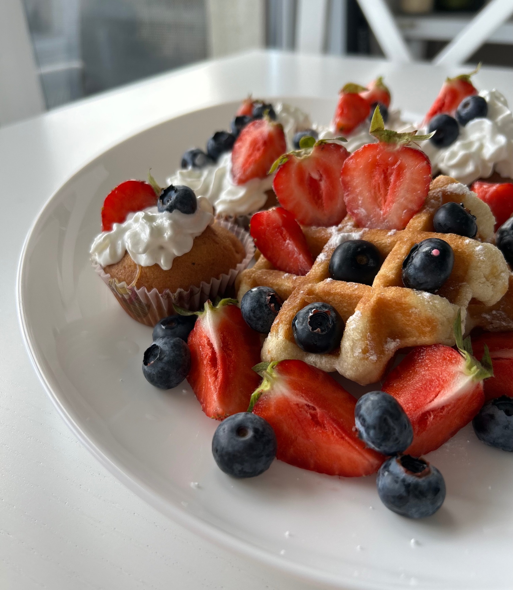 Apetyczne gofry i babeczka z bitą śmietaną, truskawkami i borówkami na białym talerzu, w jasnym otoczeniu. Słodki deser, idealny na firmowe śniadanie.