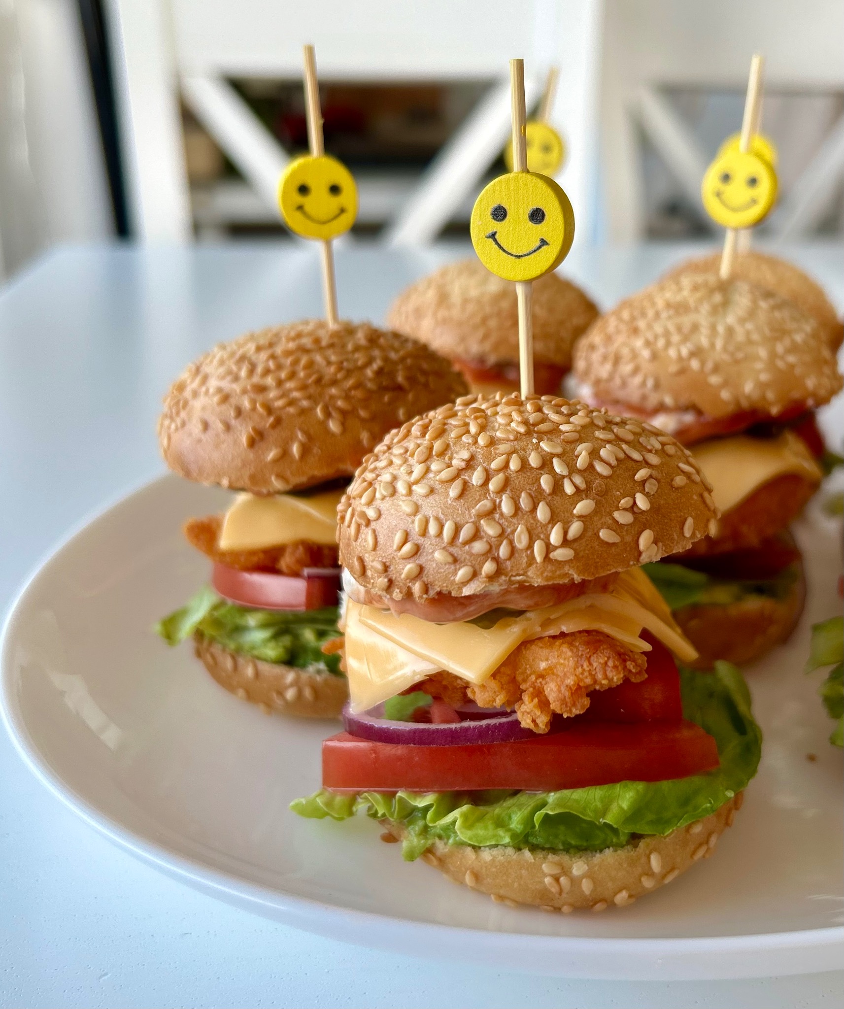 Mini burgery z serem, pomidorem i sałatą, udekorowane patyczkami z uśmiechniętymi buźkami, ułożone na białym talerzu. Jasne, naturalne oświetlenie.