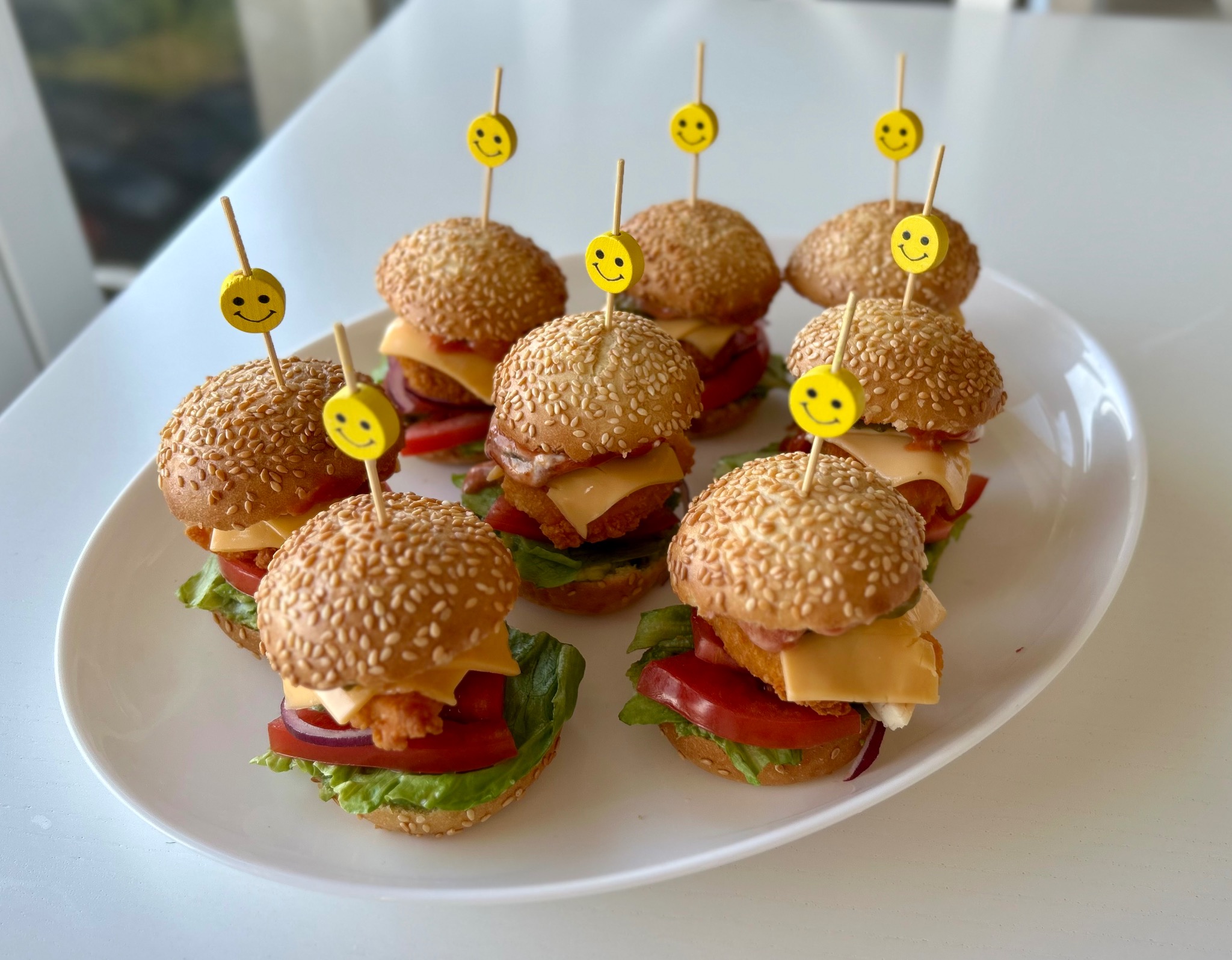 Apettyczne mini burgery z sezamową bułką i żółtymi dekoracjami uśmiechniętych buziek, ułożone na białym talerzu, idealne na przyjęcie.