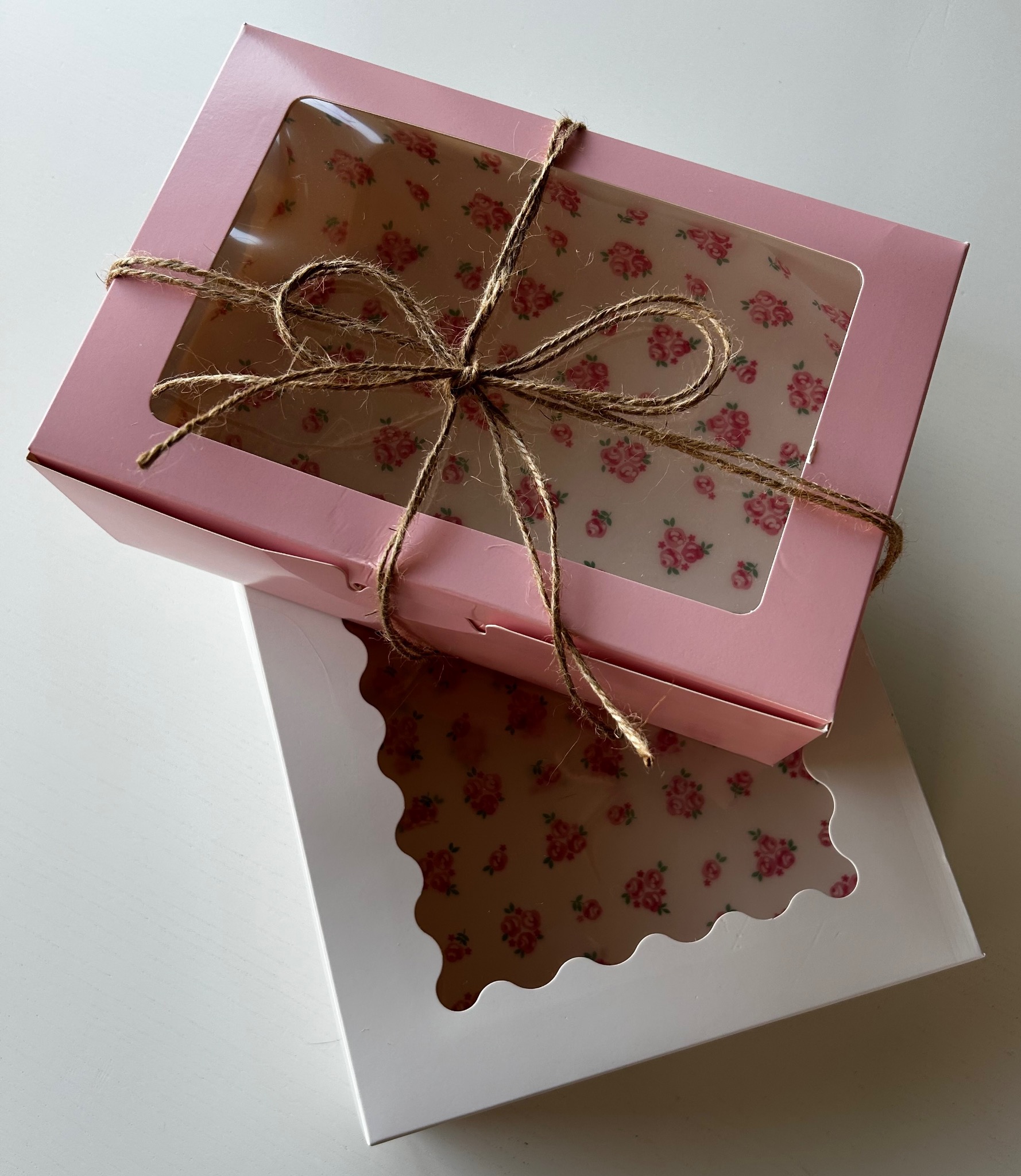 Różowe pudełko cateringowe z okienkiem, przewiązane sznurkiem, ułożone na białym pudełku z falistym brzegiem okienka, ukazujące dekoracyjne wnętrze z motywem kwiatowym.