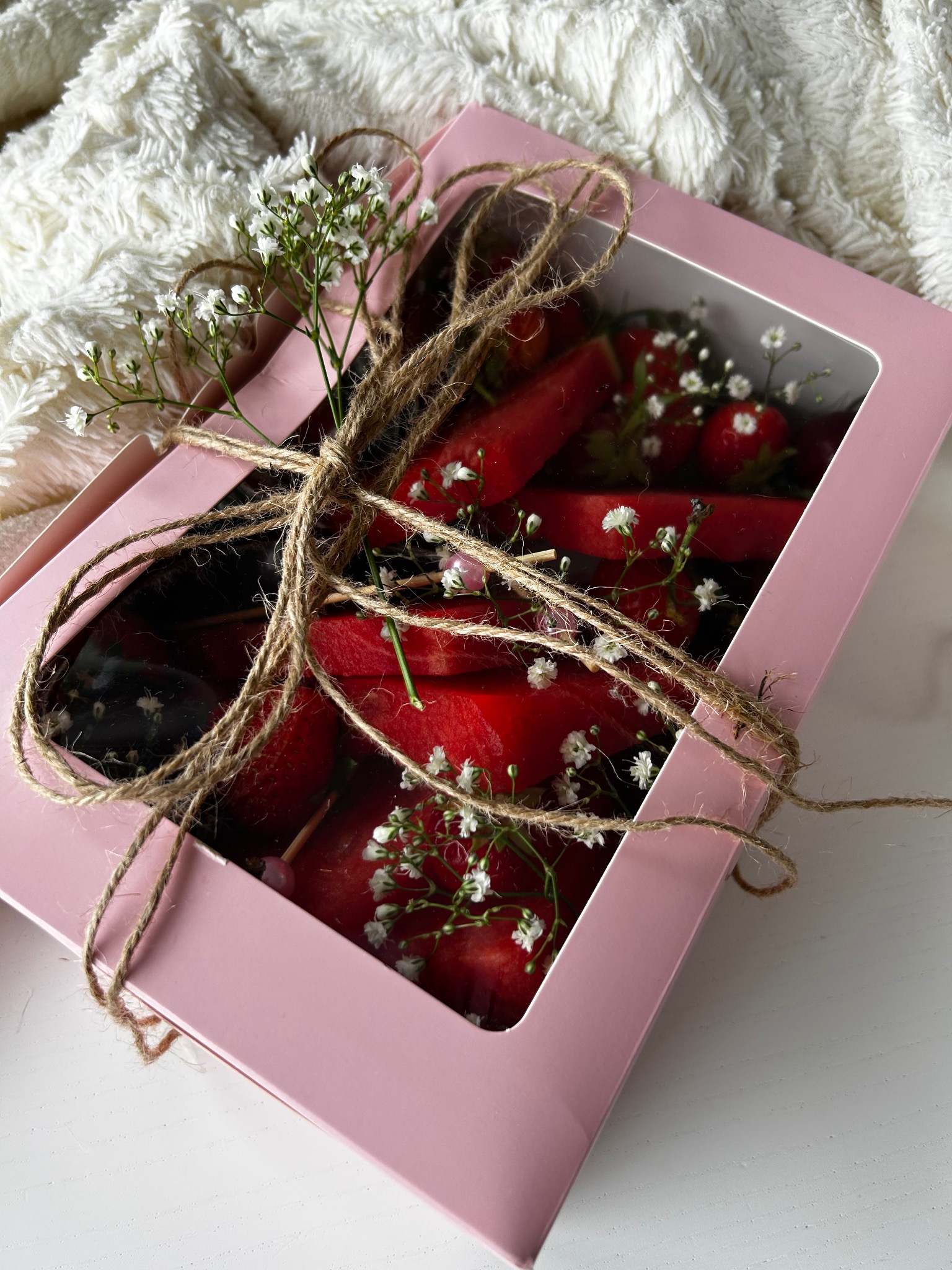 Różowe pudełko prezentowe z owocami (truskawki, arbuz) i gipsówką, przewiązane sznurkiem jutowym, na białym tle. Widok z góry na słodki upominek.