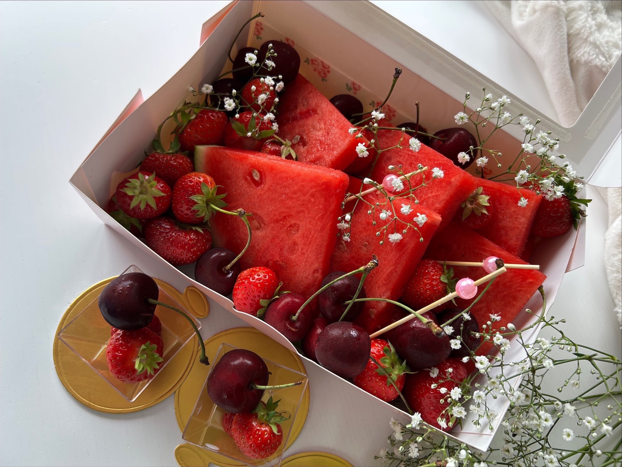 Elegancki box z kawałkami arbuza, truskawkami, czereśniami i gipsówką, na białym tle. Owoce ułożone w ozdobnym kartonowym pudełku.