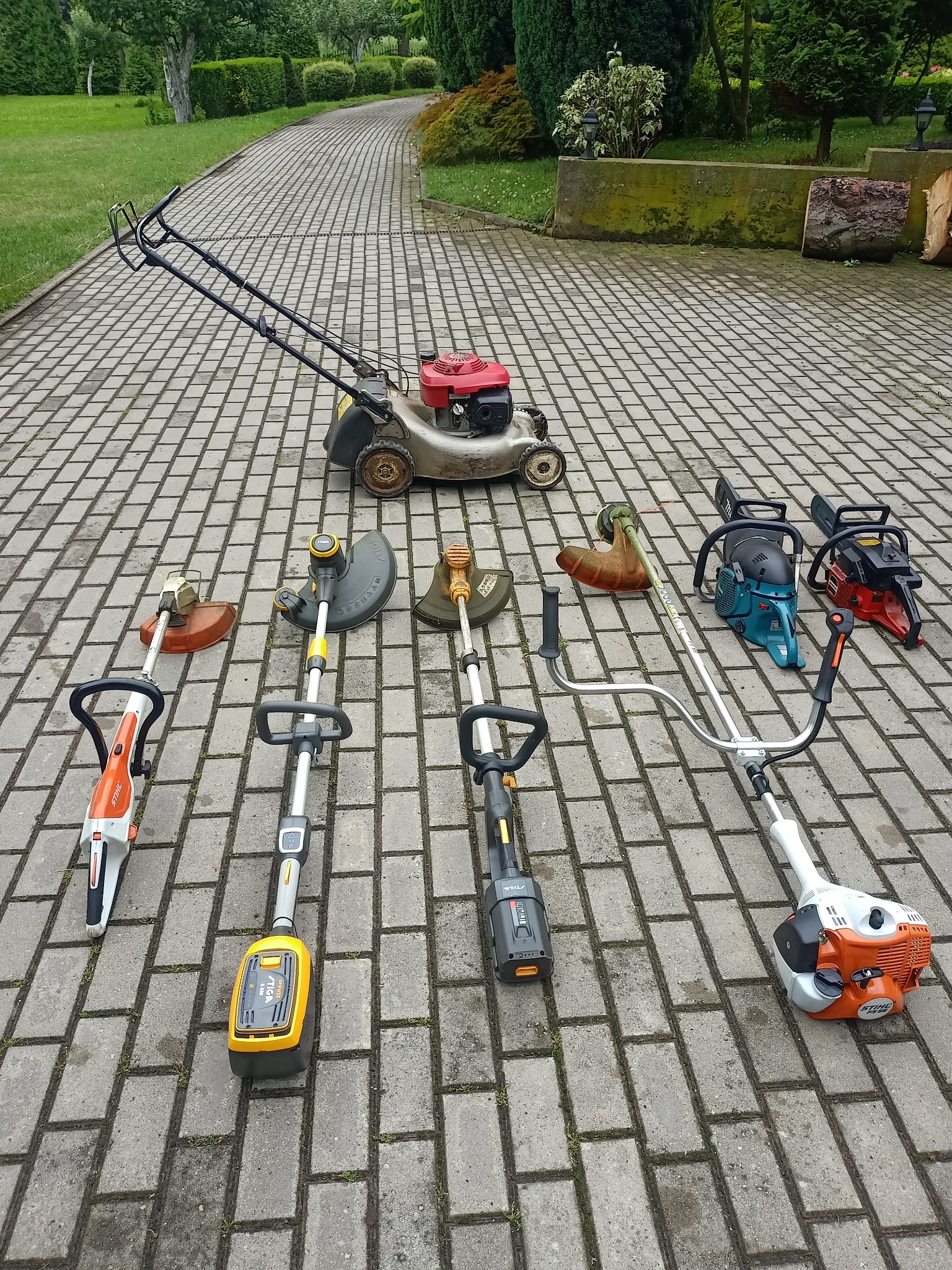Różnorodny sprzęt ogrodniczy: kosiarka, podkaszarki akumulatorowe i spalinowe, piły łańcuchowe, ułożone na brukowanym podjeździe w ogrodzie.