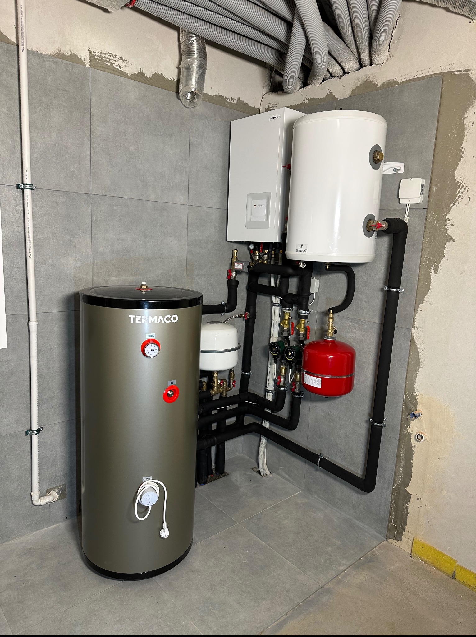 Zestaw urządzeń grzewczych: bojler Termaco, biały zasobnik Galmet i piec Hitachi, połączone czarnymi rurami w kotłowni wykończonej szarą płytką. Widoczne rury pod sufitem.