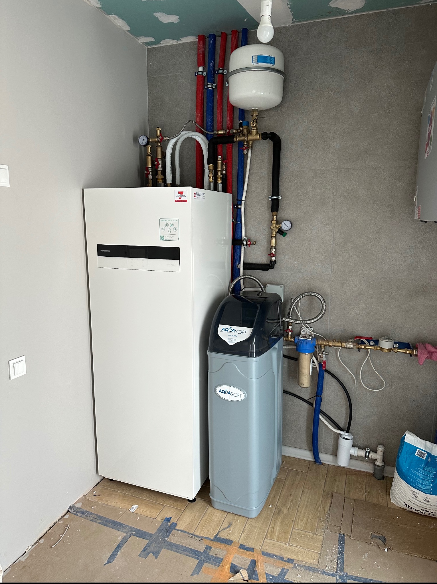 Biała pompa ciepła Panasonic Aquarea, obok zmiękczacz wody Aquasoft, rury instalacyjne w kolorach niebieskim i czerwonym, manometry, szary gres na ścianie.