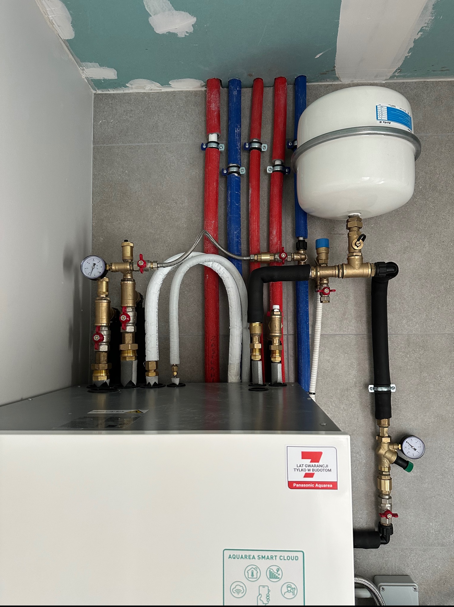 Instalacja pompy ciepła Panasonic Aquarea Smart Cloud z rurami w kolorach czerwonym i niebieskim, zbiornikiem wyrównawczym i manometrami, zamontowana na szarej ścianie.