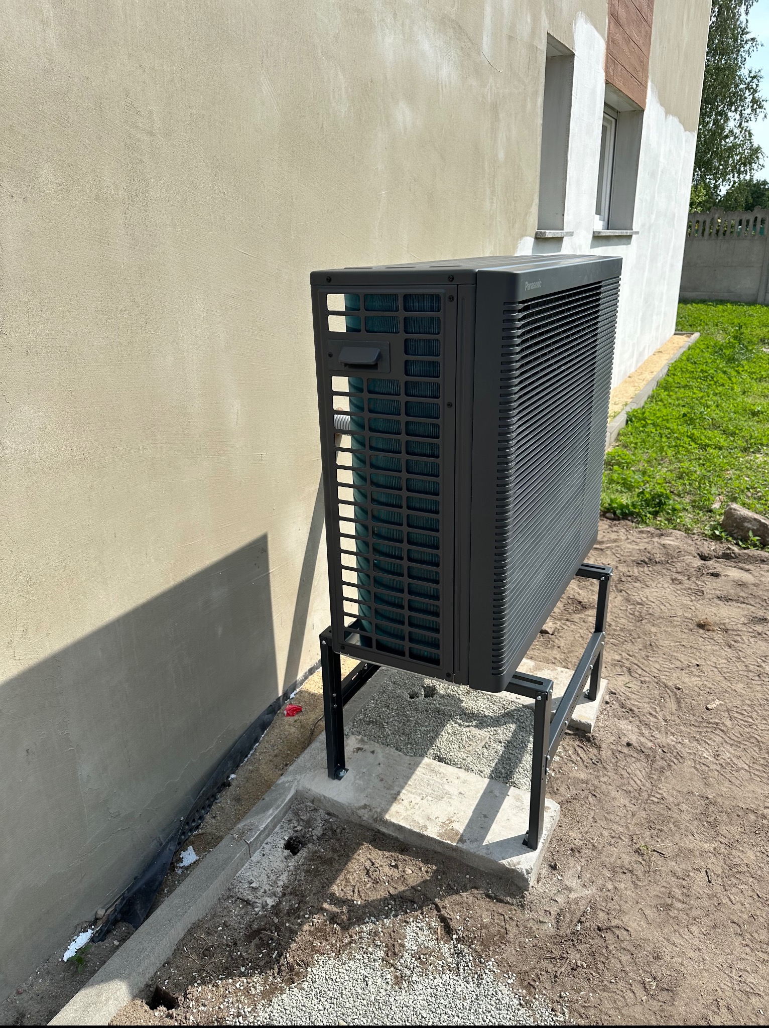 Zewnętrzna jednostka pompy ciepła Panasonic, zamontowana na metalowej konstrukcji przy ścianie budynku w trakcie wykończenia. Widoczne elementy instalacji i otoczenie.