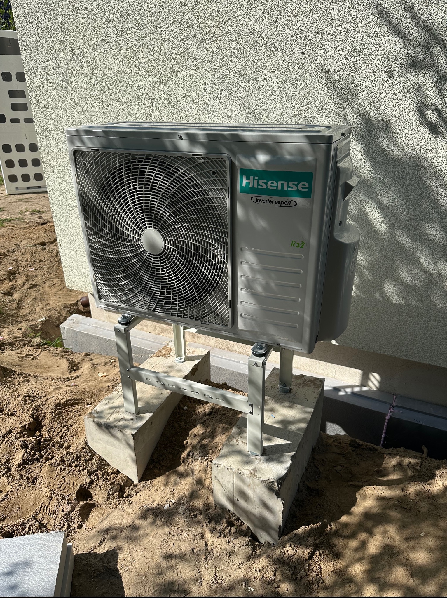 Zamontowana jednostka zewnętrzna klimatyzacji Hisense na betonowych podstawach przy ścianie budynku z fakturą. Widoczne ślady prac ziemnych.