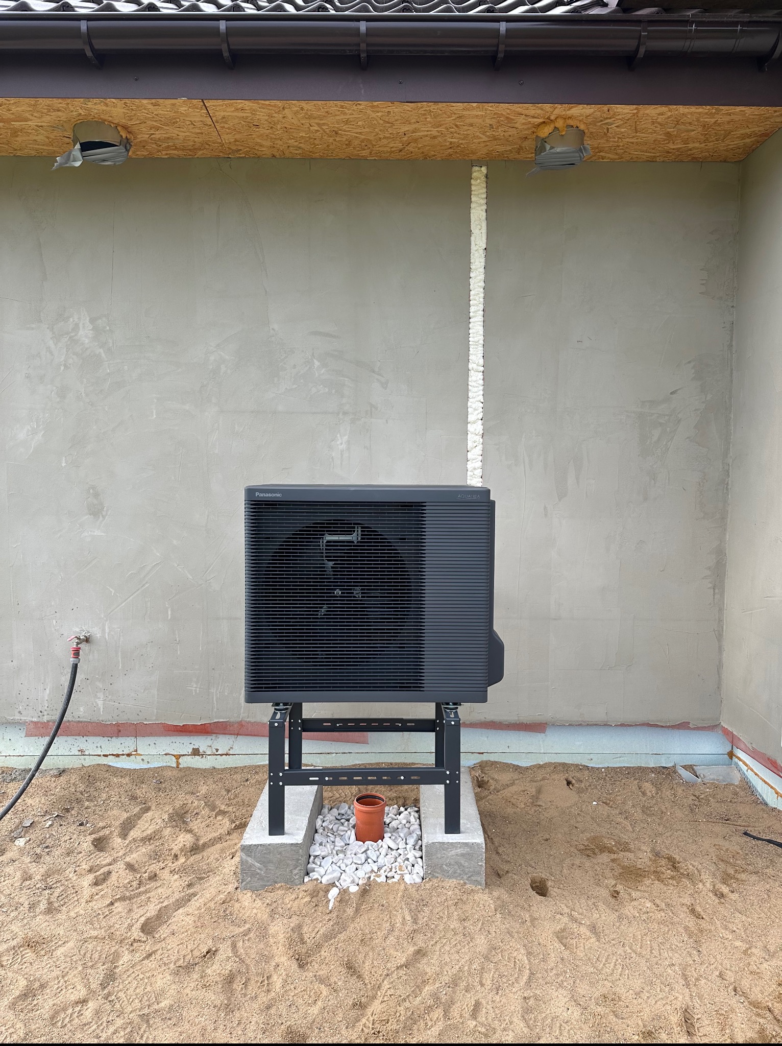 Zewnętrzna jednostka pompy ciepła Panasonic Aquarea, zamontowana na betonowym fundamencie z białym kamieniem, na tle szarej ściany budynku w trakcie prac wykończeniowych.