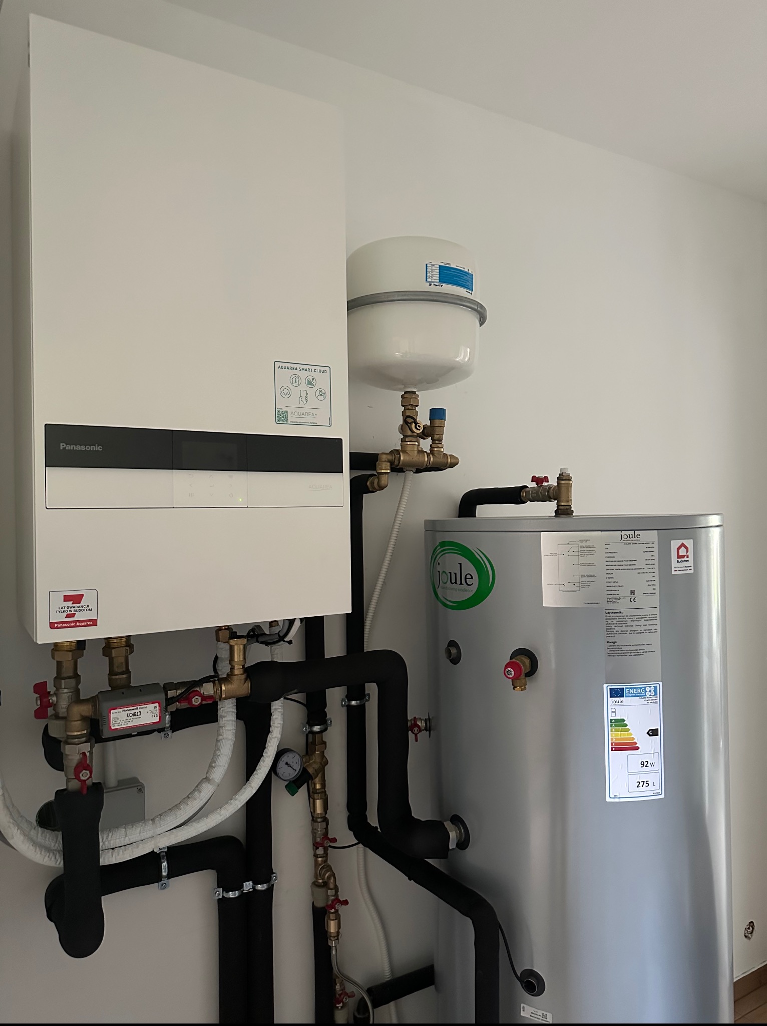 Biała pompa ciepła Panasonic Aquarea z szarym zasobnikiem Joule, instalacja z czarnych rur. Widoczne naklejki informacyjne i oznaczenia energetyczne.