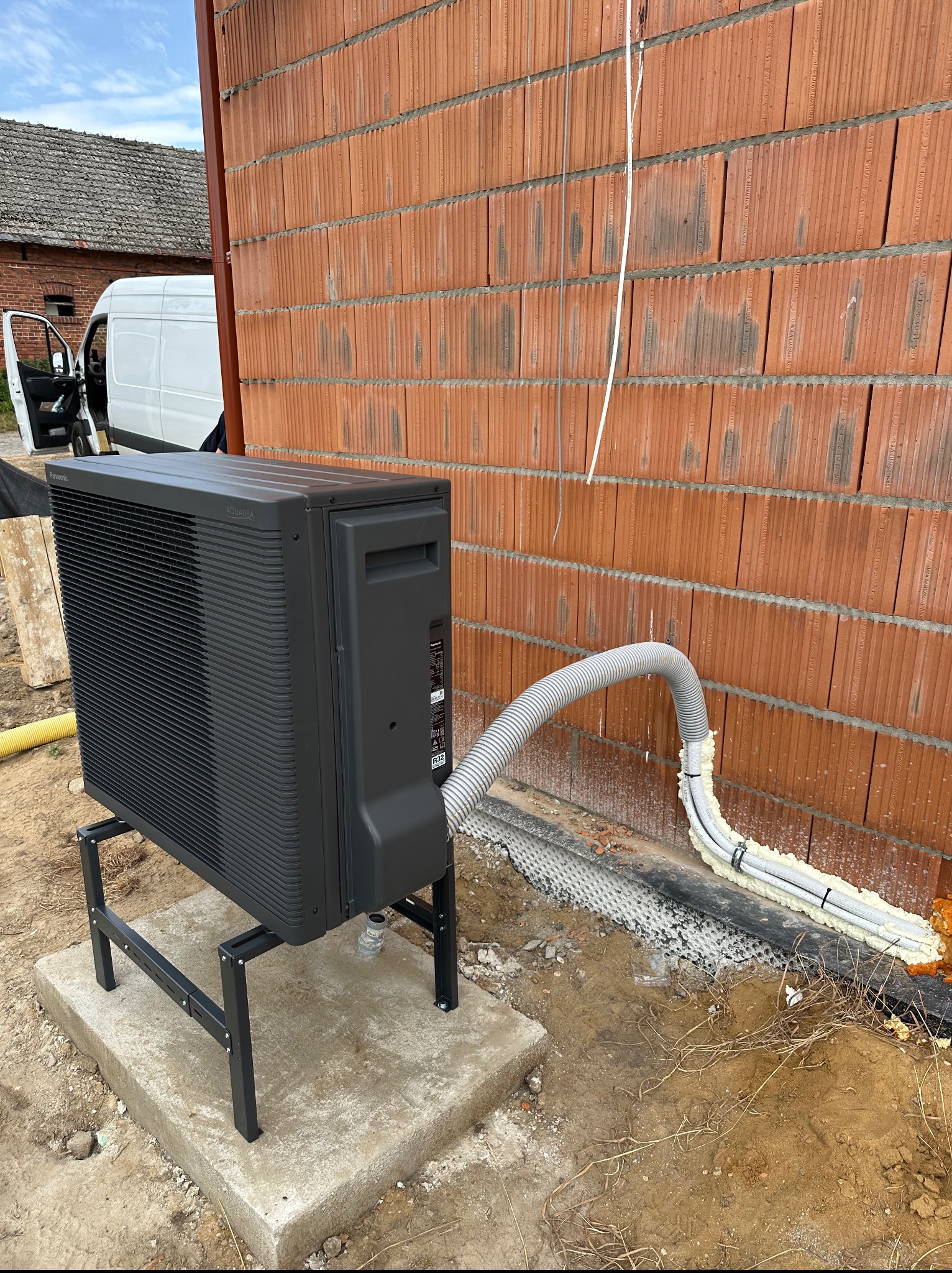 Czarna pompa ciepła Panasonic Aquarea zamontowana przy ceglanej ścianie budynku w trakcie instalacji. Widoczne rury i przewody. Biały van w tle.
