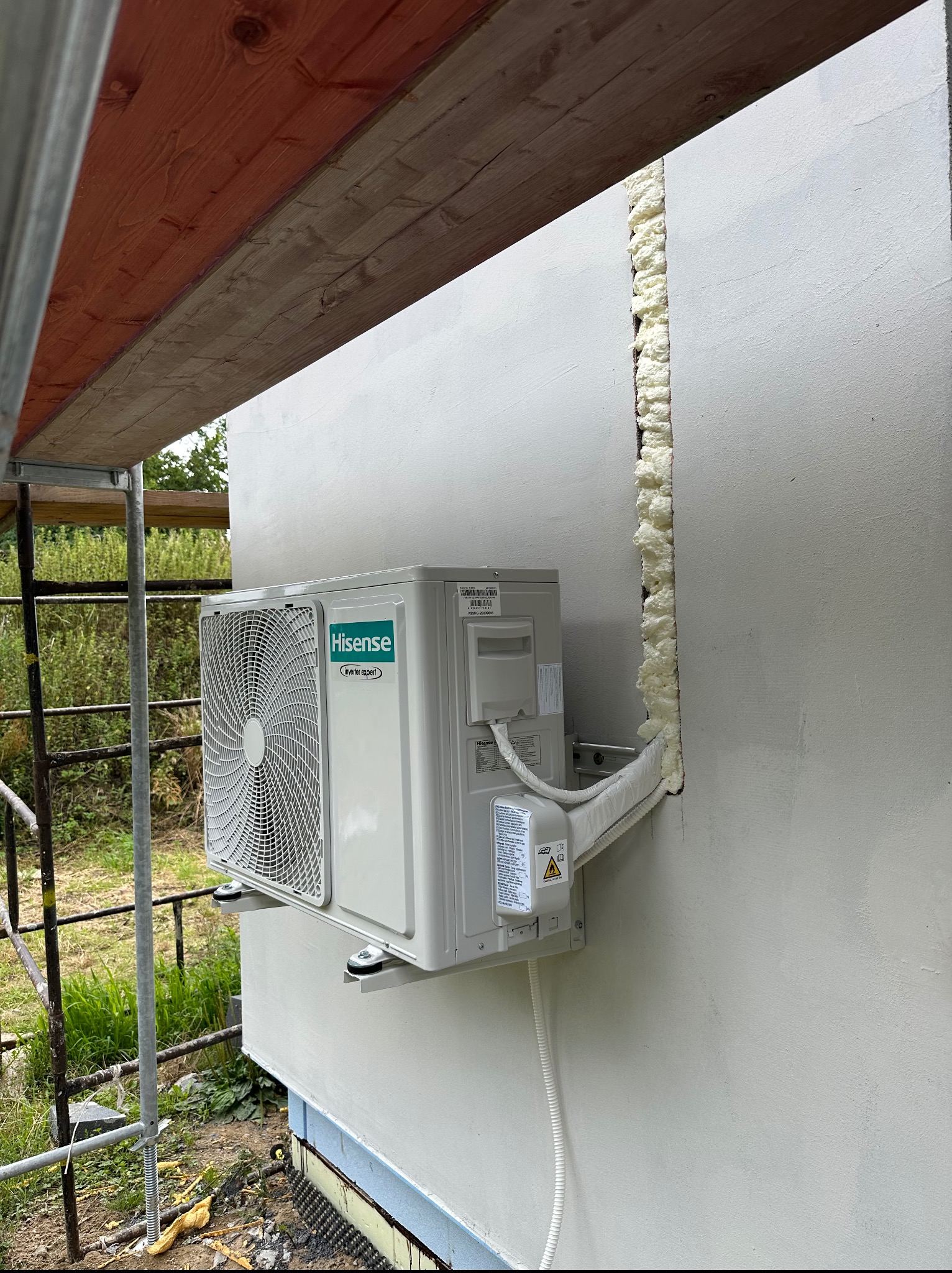 Biała jednostka zewnętrzna klimatyzacji marki Hisense zamontowana na ścianie budynku z widoczną pianką montażową w szczelinie. Widoczne rusztowanie.