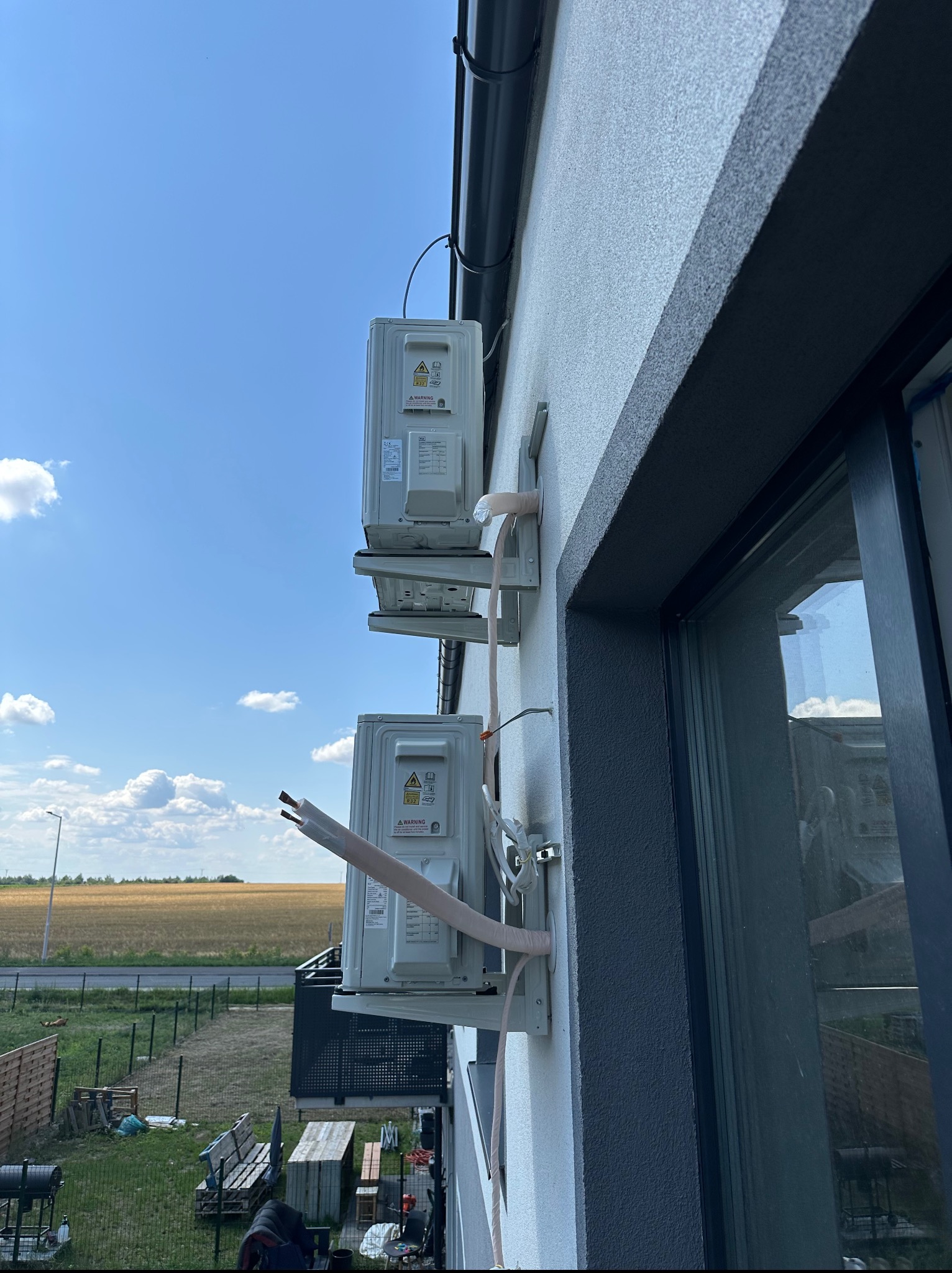 Dwa klimatyzatory zamontowane na elewacji domu, widoczne przewody instalacyjne. W tle błękitne niebo i pole. Kąt ujęcia z dołu.