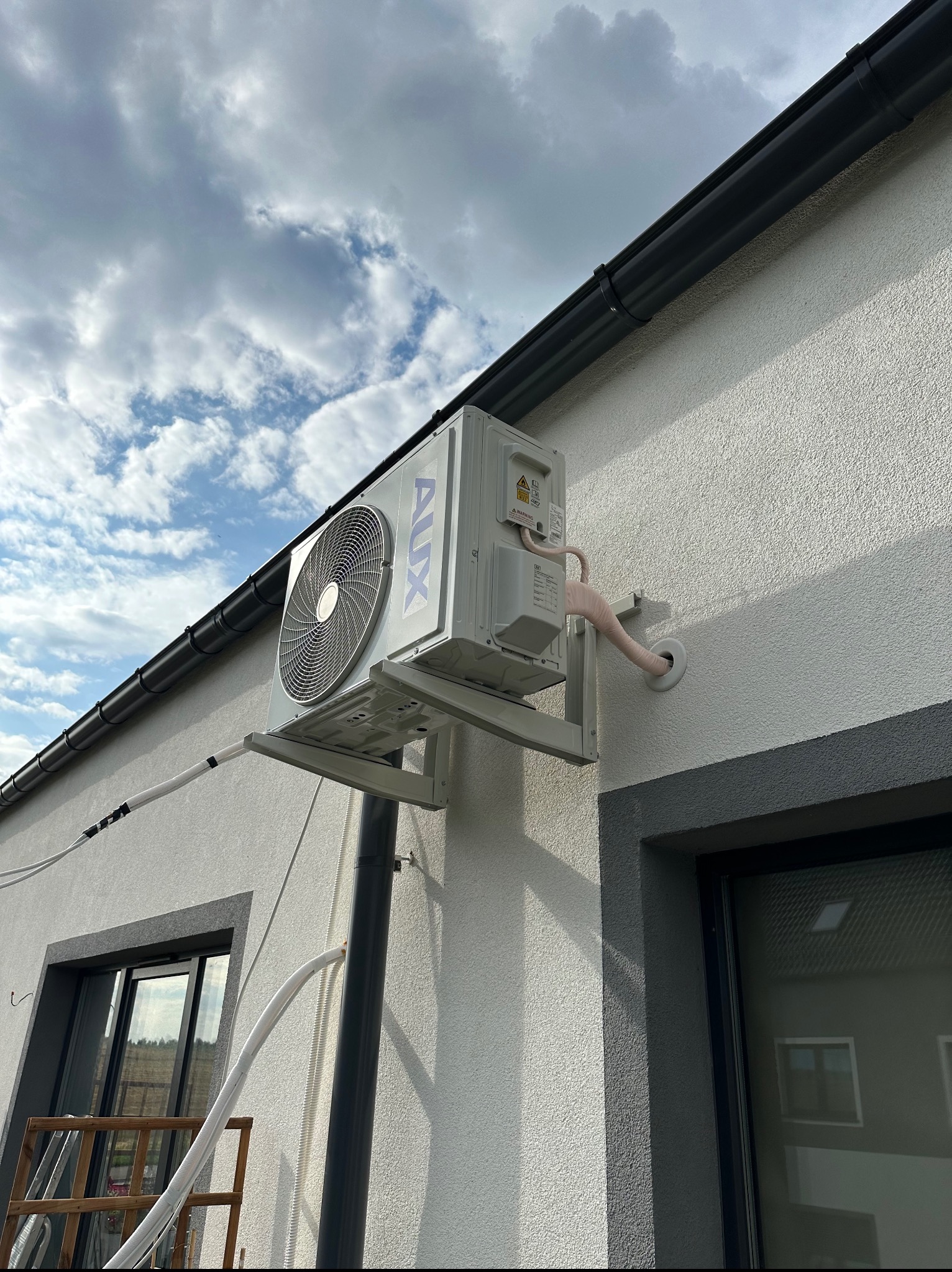 Biała jednostka zewnętrzna klimatyzacji AUX zamontowana na elewacji budynku z widocznymi przewodami i elementami instalacji. Tło stanowi pochmurne niebo.