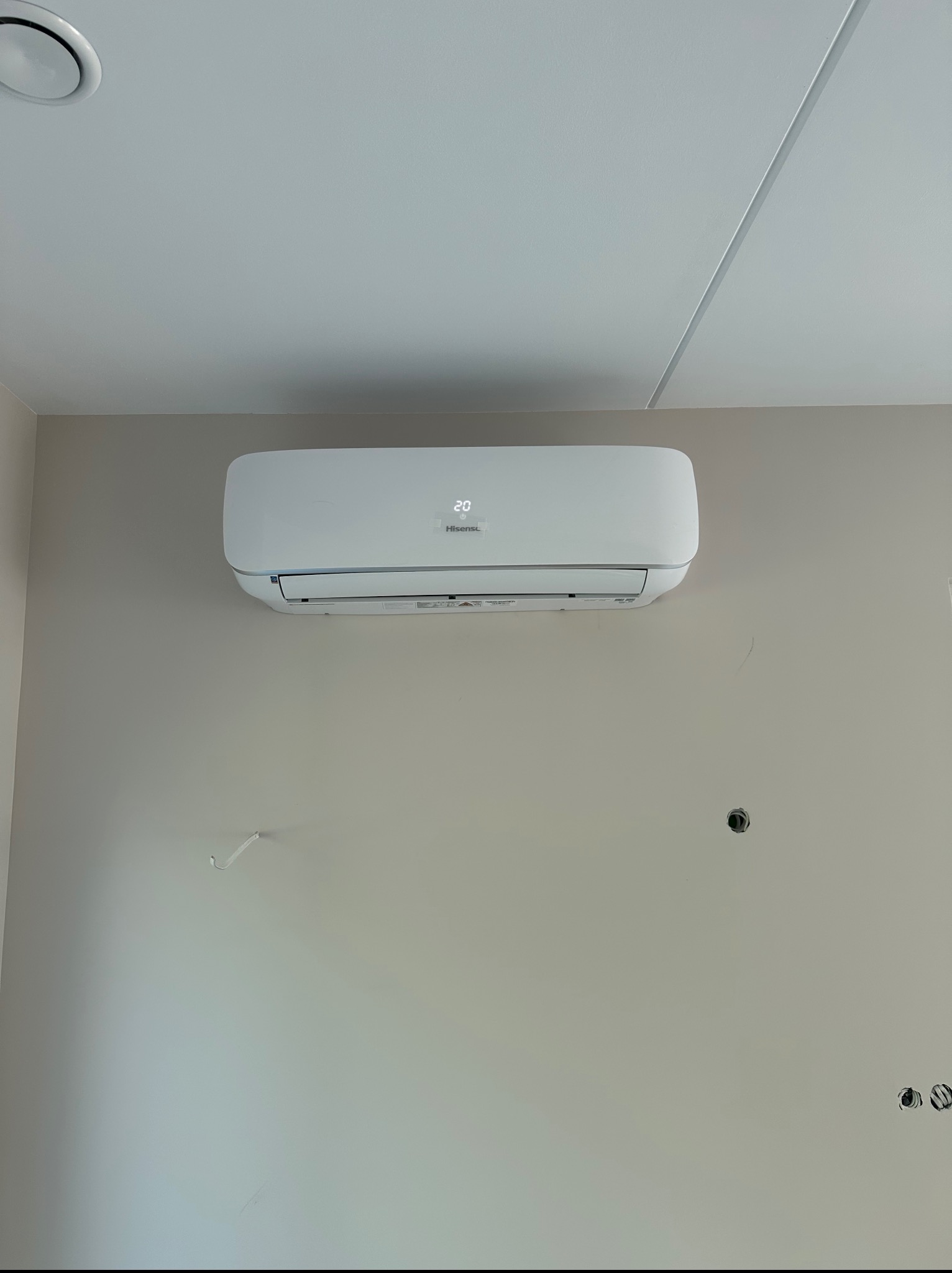 Biała klimatyzacja Hisense zamontowana na beżowej ścianie w pomieszczeniu. Widoczny wyświetlacz z temperaturą 20 stopni. Na ścianie ślady po instalacji.