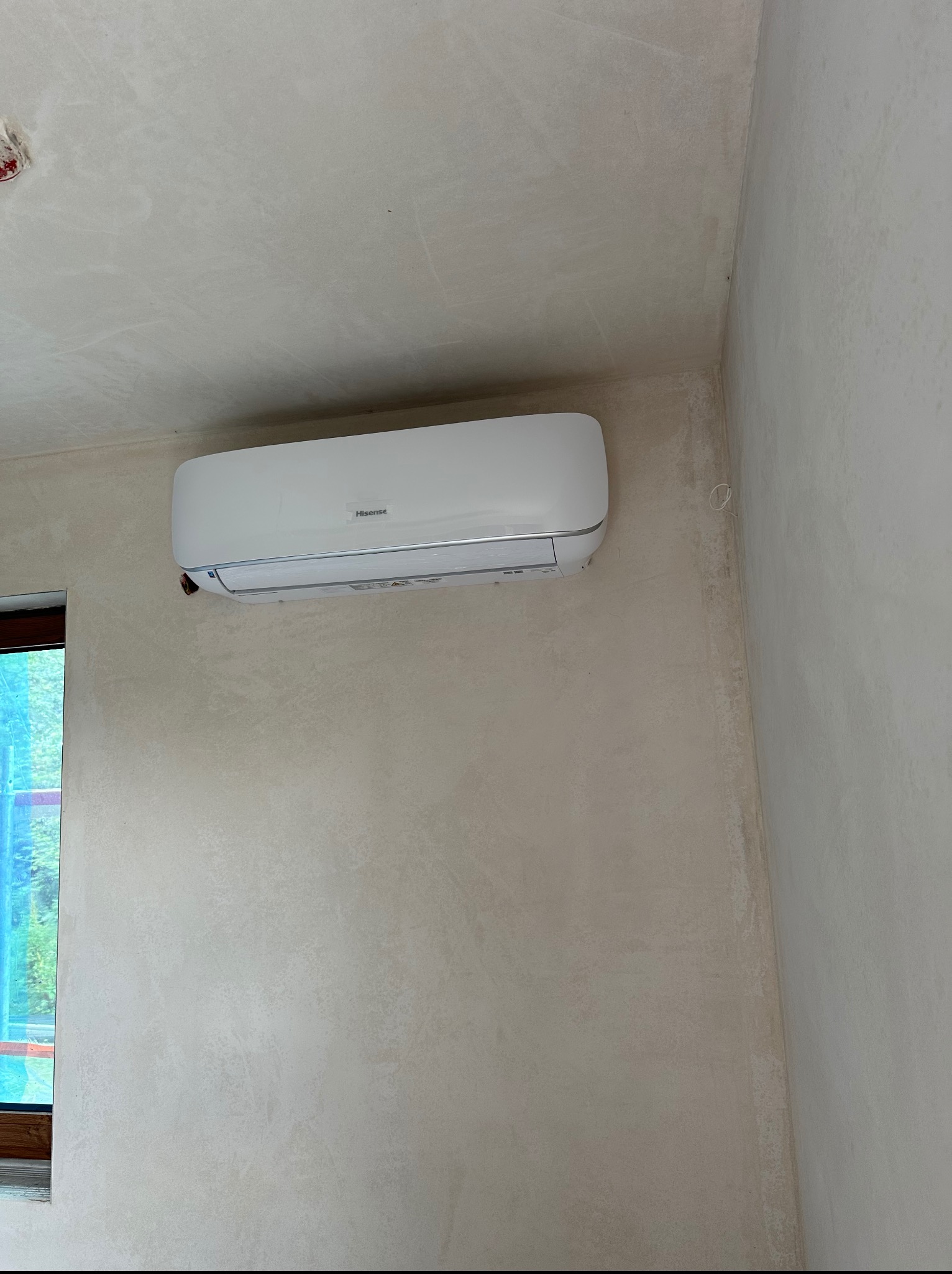 Biały klimatyzator Hisense zamontowany na ścianie o fakturze betonu w rogu pokoju, widoczne okno z boku. Nowoczesny design, minimalistyczne wnętrze.