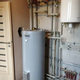 Szary, pionowy bojler Ferroli z podłączeniami hydraulicznymi i narzędziami na górze, umieszczony w pomieszczeniu z widoczną instalacją grzewczą i drzwiami po lewej.