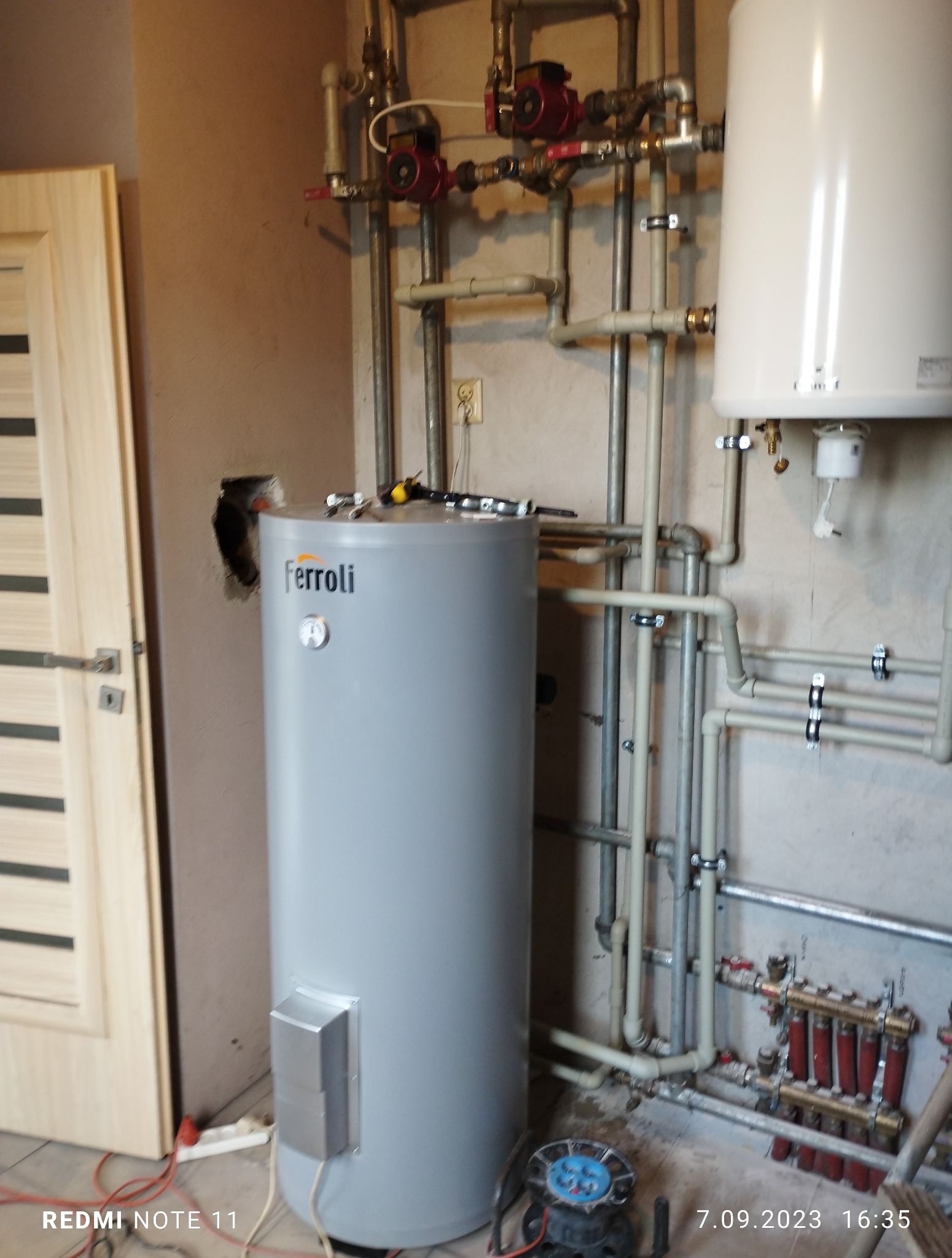 Szary, pionowy bojler Ferroli z podłączeniami hydraulicznymi i narzędziami na górze, umieszczony w pomieszczeniu z widoczną instalacją grzewczą i drzwiami po lewej.