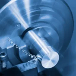 Metalowy wałek obrabiany na tokarce z wiórami, w niebieskiej tonacji. Widoczny fragment tokarki i śruba mocująca. Obróbka skrawaniem, zbliżenie.