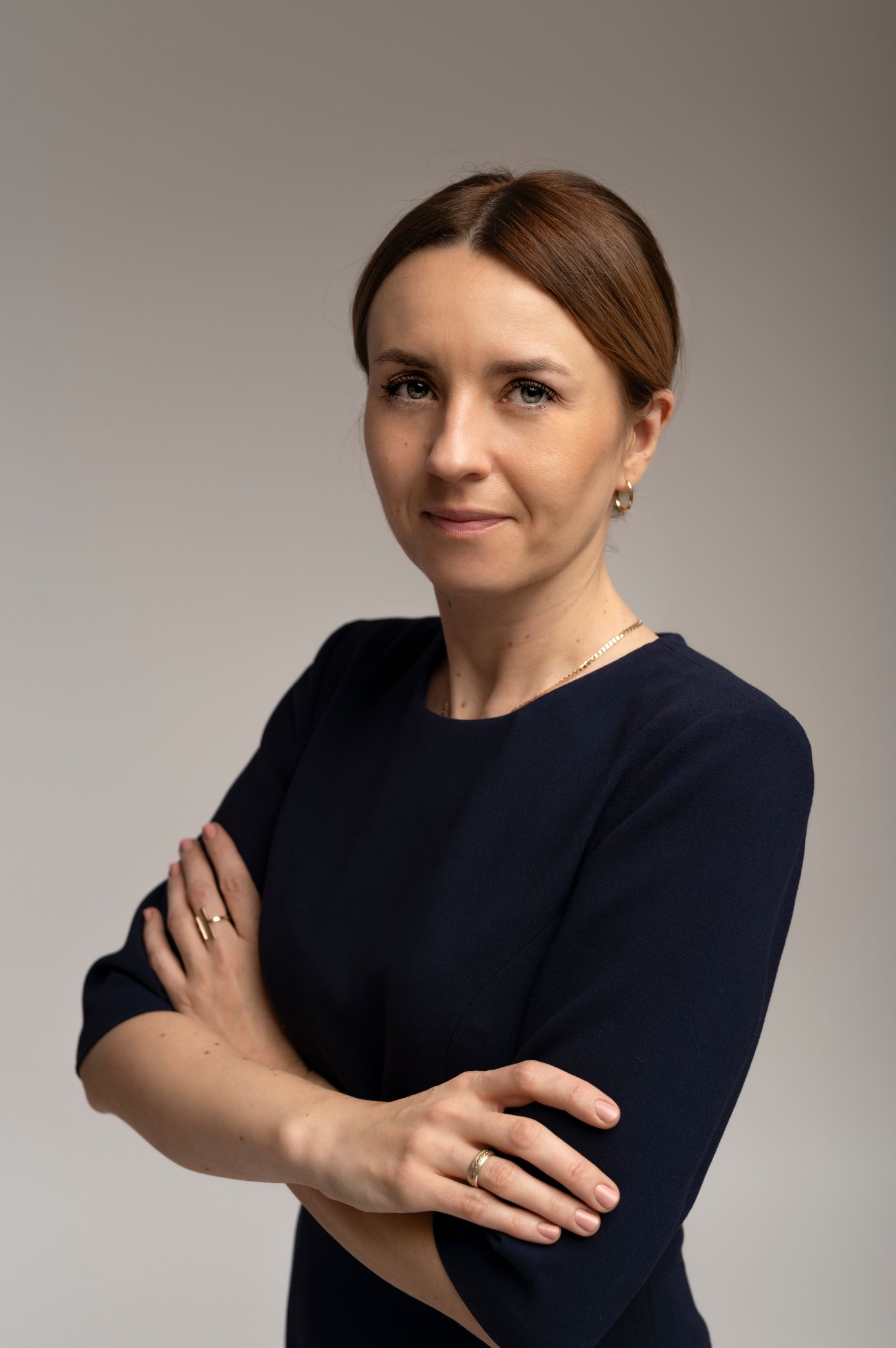 Portret kobiety w granatowej sukience, z rękami skrzyżowanymi na piersiach, na neutralnym tle. Widoczne detale biżuterii: złoty łańcuszek i pierścionki.