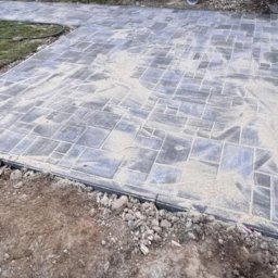 Lucas Stones - Ułożona kostka brukowa w geometryczny wzór, zasypana piaskiem, z widocznym obrzeżem i fragmentem trawnika. Częściowo ukończona nawierzchnia.
