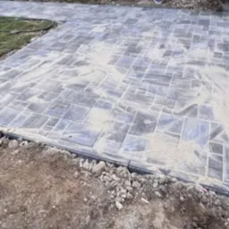 Ułożona kostka brukowa w geometryczny wzór, zasypana piaskiem, z widocznym obrzeżem i fragmentem trawnika. Częściowo ukończona nawierzchnia.