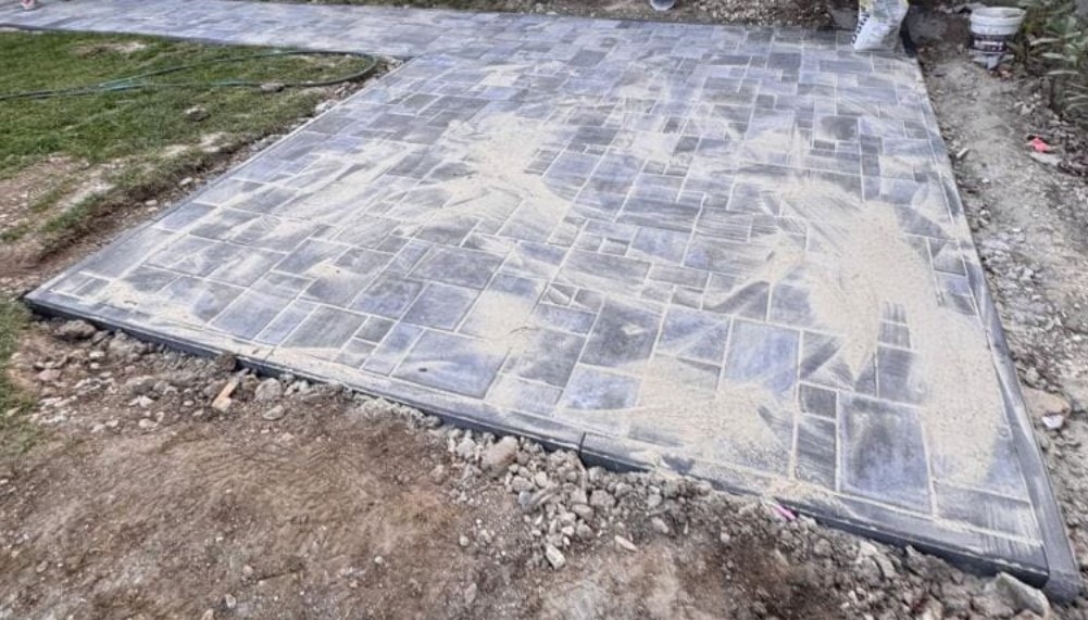 Ułożona kostka brukowa w geometryczny wzór, zasypana piaskiem, z widocznym obrzeżem i fragmentem trawnika. Częściowo ukończona nawierzchnia.