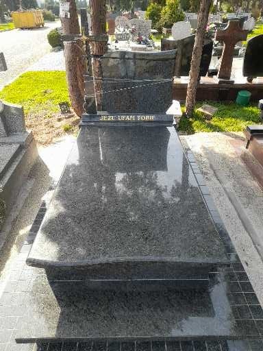 Polerowany, ciemny nagrobek z napisem 'Jezu Ufam Tobie' na cmentarzu, otoczony zielenią i innymi nagrobkami w tle. Widok z góry.