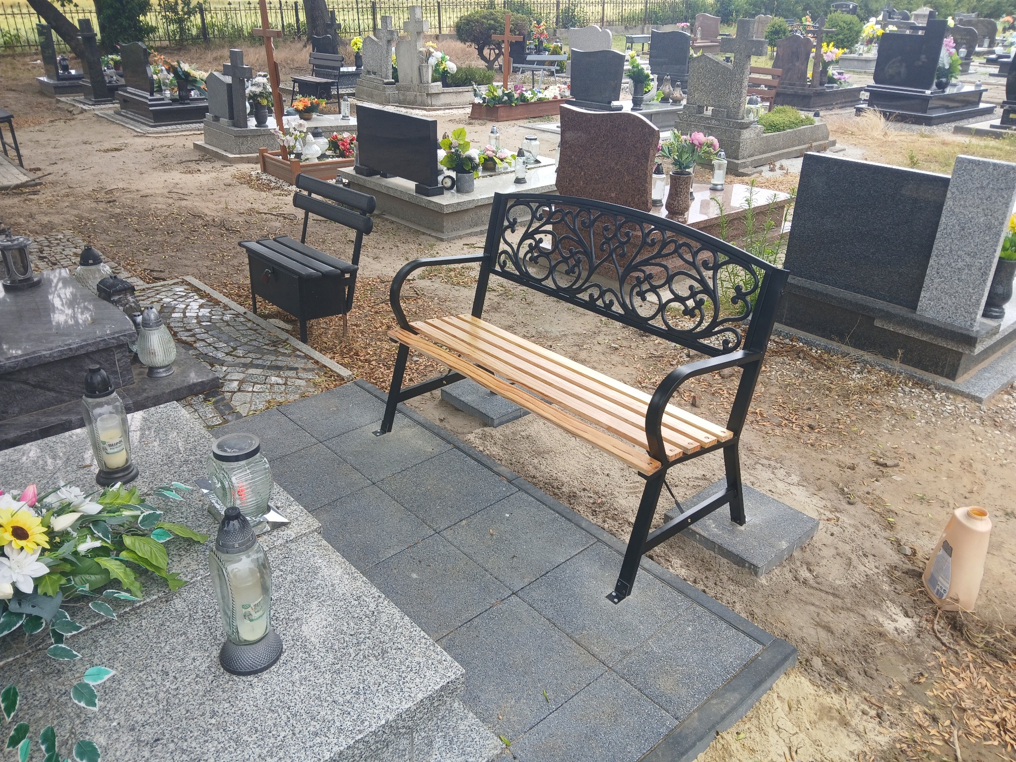 Cmentarz: ławka z drewnianym siedziskiem i metalowym oparciem, ułożona na szarych płytach chodnikowych, wśród nagrobków i zniczy. Widok ogólny.