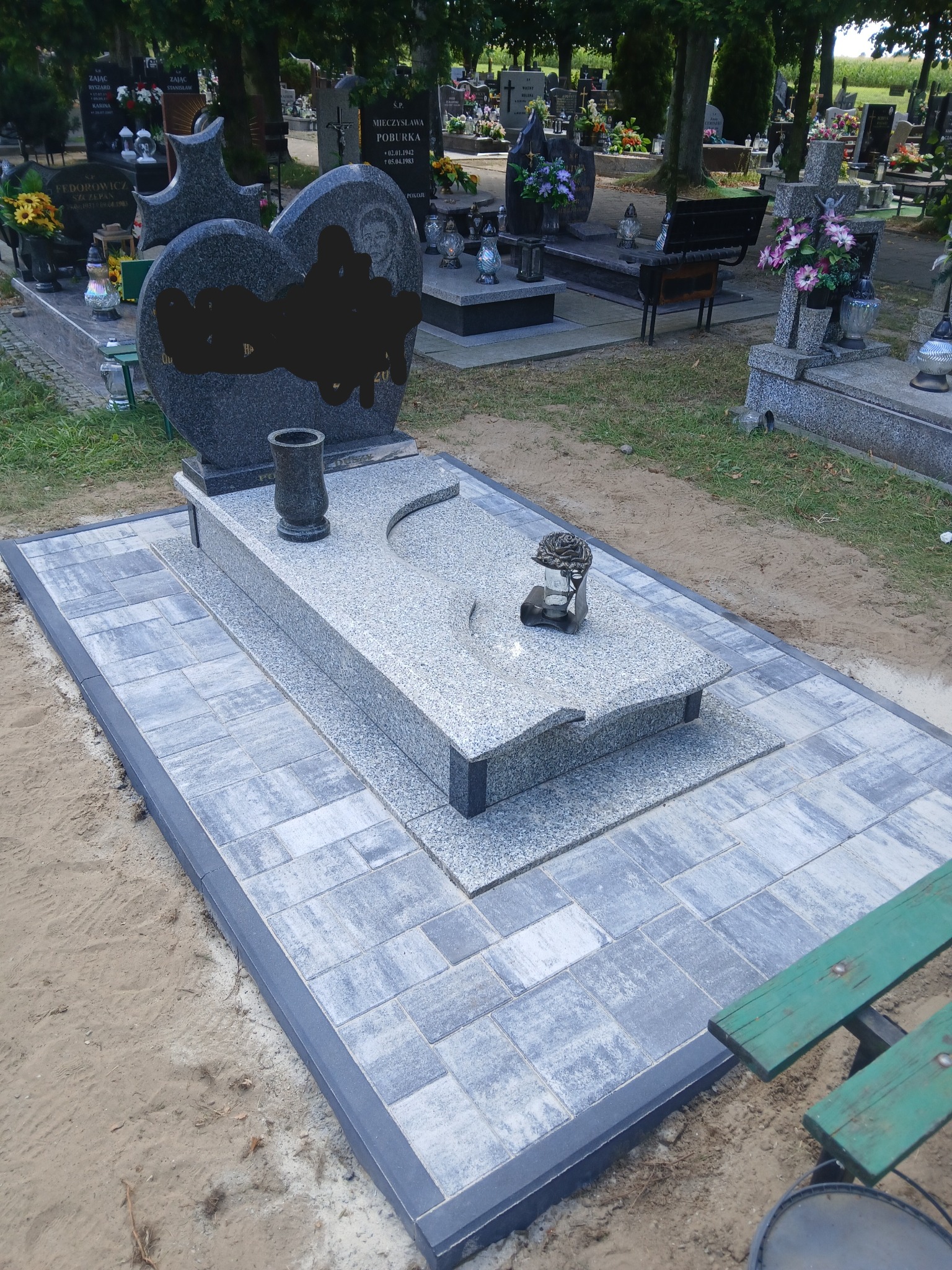 Granitowy nagrobek z szarą kostką brukową na cmentarzu, z widoczną urną i zniczem w kształcie róży. Nagrobek w kształcie serca z czarnym elementem.