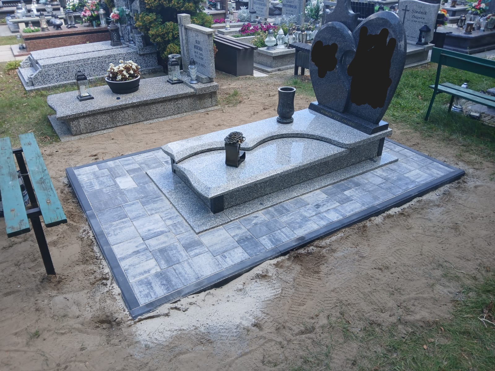 Granitowy nagrobek z sercami i wazonem, otoczony nową kostką brukową na cmentarzu. Widoczne inne nagrobki i ławki w tle. Jasne, naturalne oświetlenie.