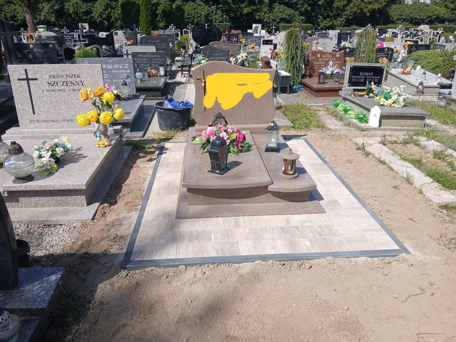 Nowy nagrobek z jasnego kamienia z kwiatami i lampionami, otoczony świeżo ułożoną kostką brukową na cmentarzu w słoneczny dzień. Widoczne inne groby w tle.