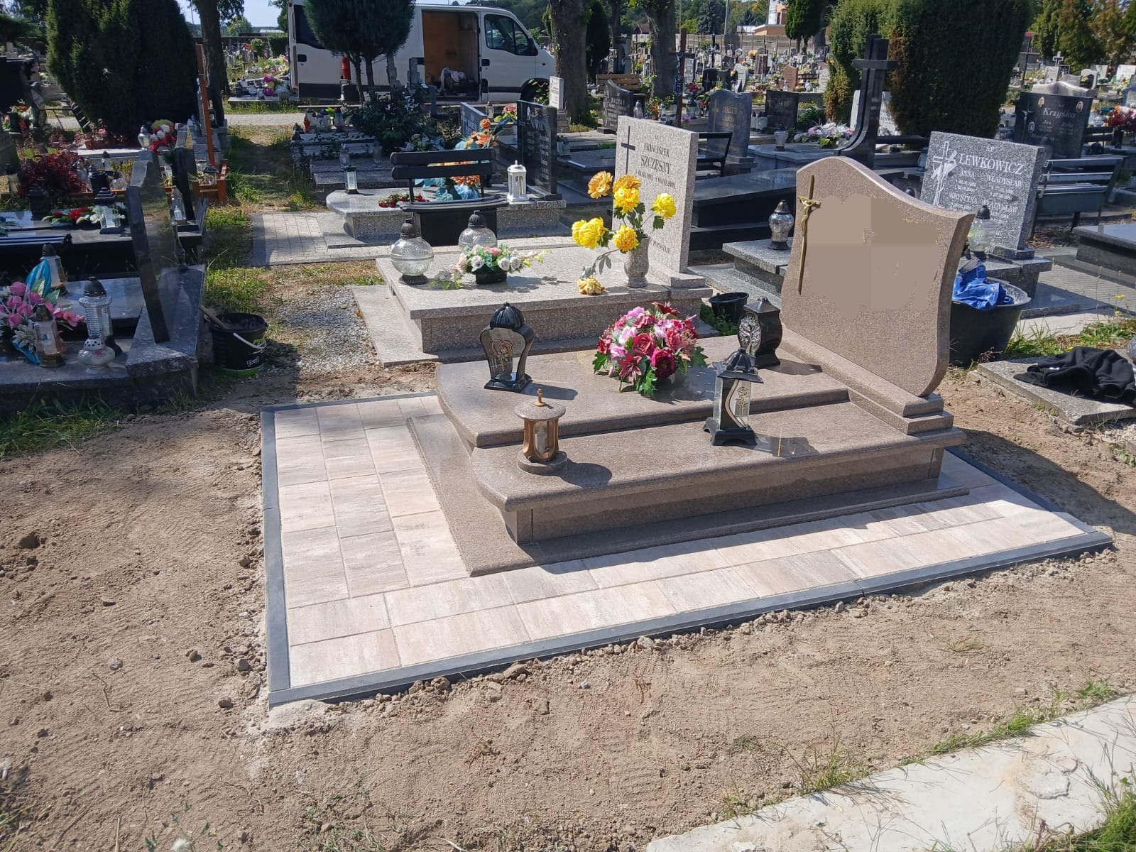 Jasny nagrobek z dekoracjami kwiatowymi i lampionami, otoczony świeżo ułożoną kostką brukową na cmentarzu w słoneczny dzień.