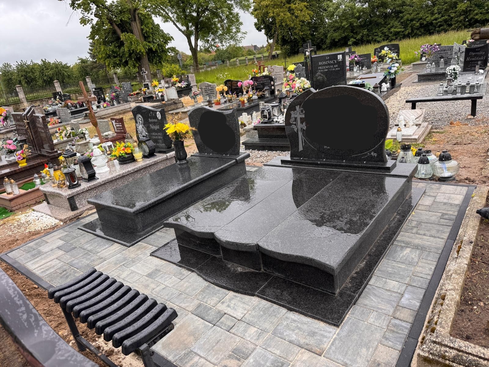 Granitowy nagrobek z czarnym, błyszczącym kamieniem i ławką na cmentarzu w Brzegu. Otoczenie wyłożone kostką brukową. Widok na inne groby.